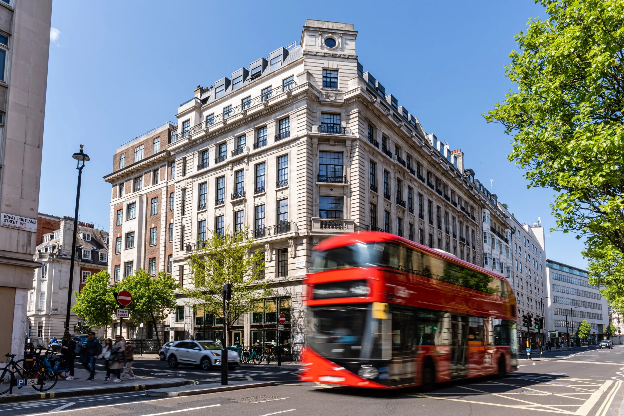 Red double-decker bus moving across a city street with pedestrians walking and cars parked, buildings in the background with Crittall windows, and trees lining the street on a sunny day.