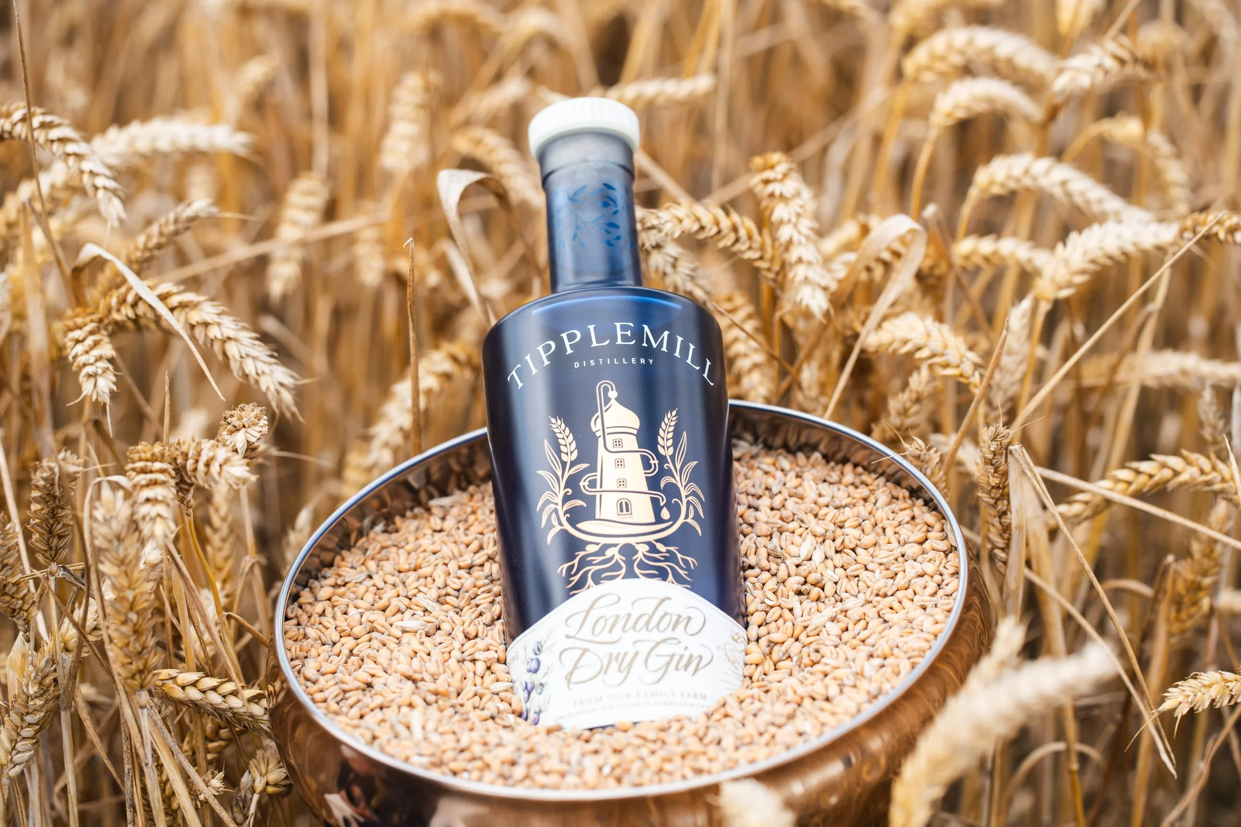 A bottle of Tippelmill London Dry Gin sitting in a metal container filled with wheat grains, inside a wheat field.