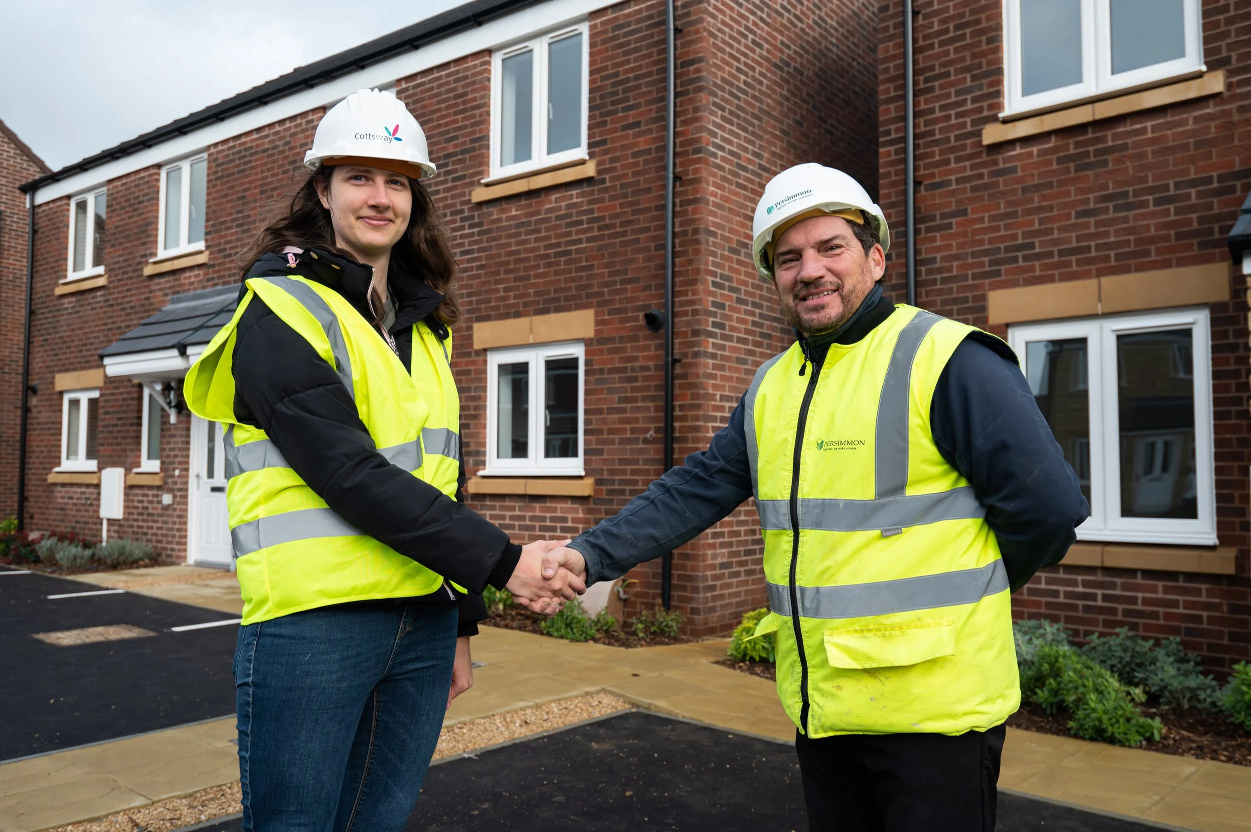 Two construction workers shaking hands in front of a residential building, both wearing high-visibility yellow vests and white safety helmets.