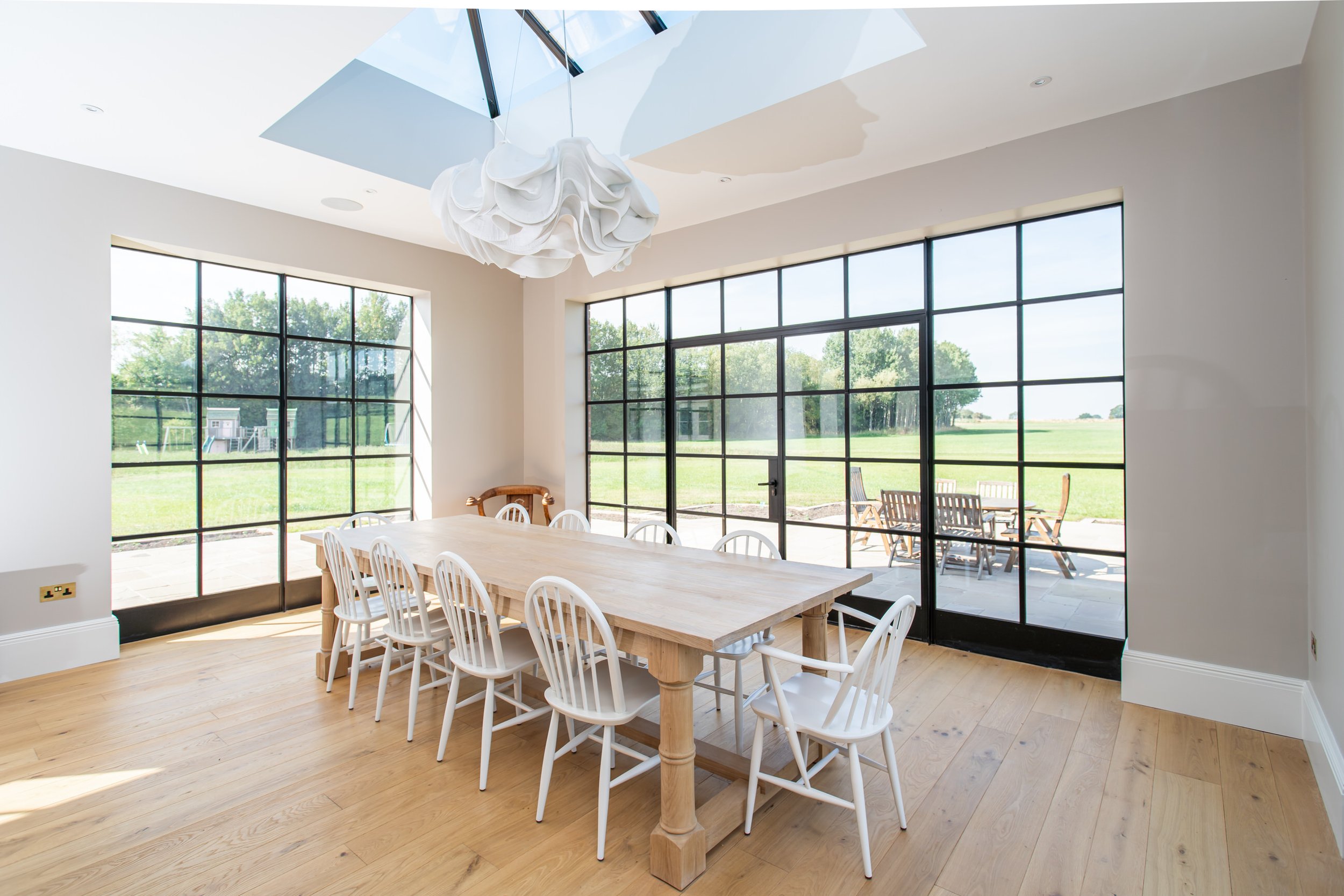 Bright dining room with large black-framed Crittall windows and sliding glass door, light wood dining table with white chairs, wooden outdoor furniture on patio, skylight, light-colored walls, hardwood floor.