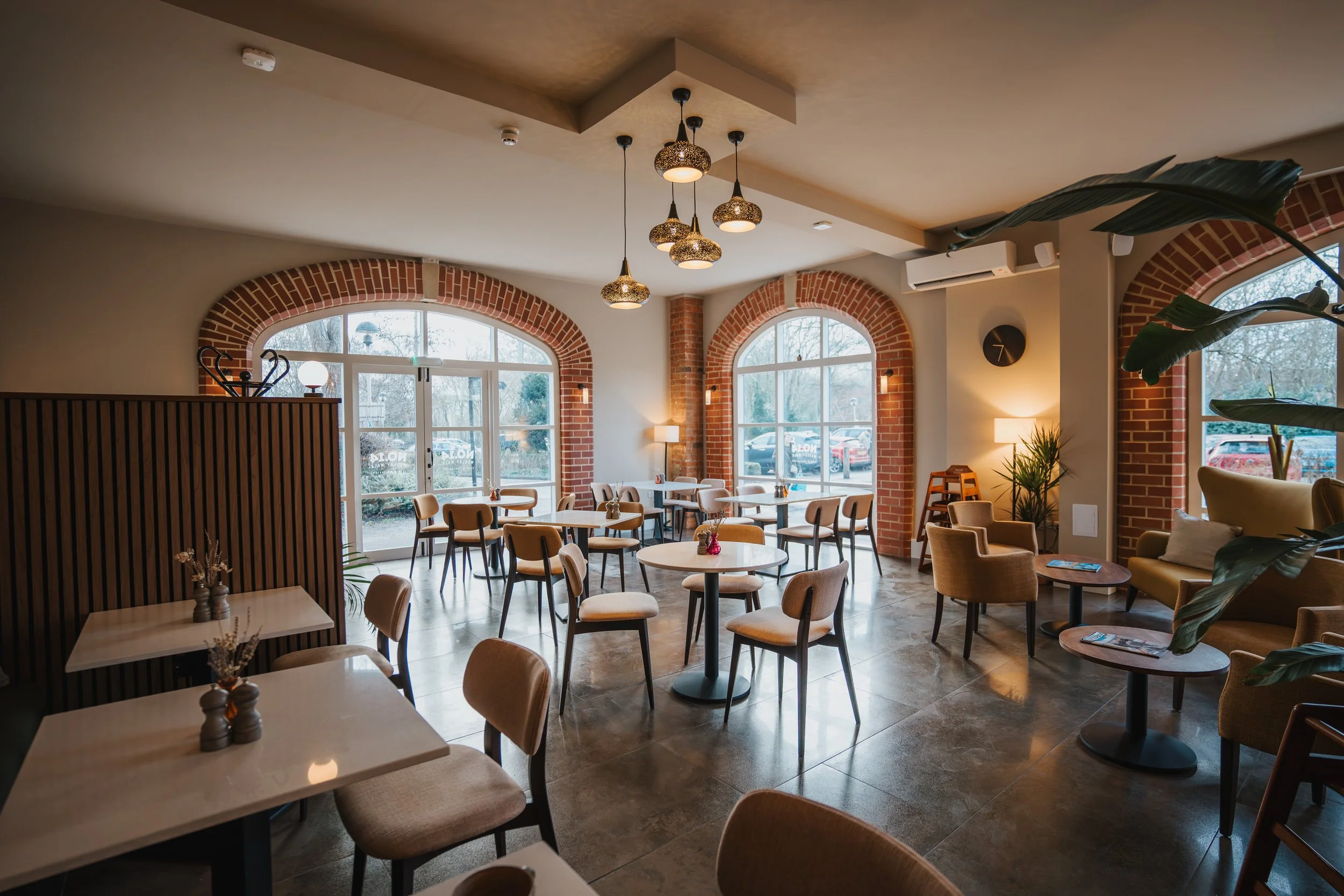 Cozy restaurant interior with large arched windows, wooden chairs, round tables with small vases, hanging pendant lights, and a mix of seating options, including armchairs and a sofa.