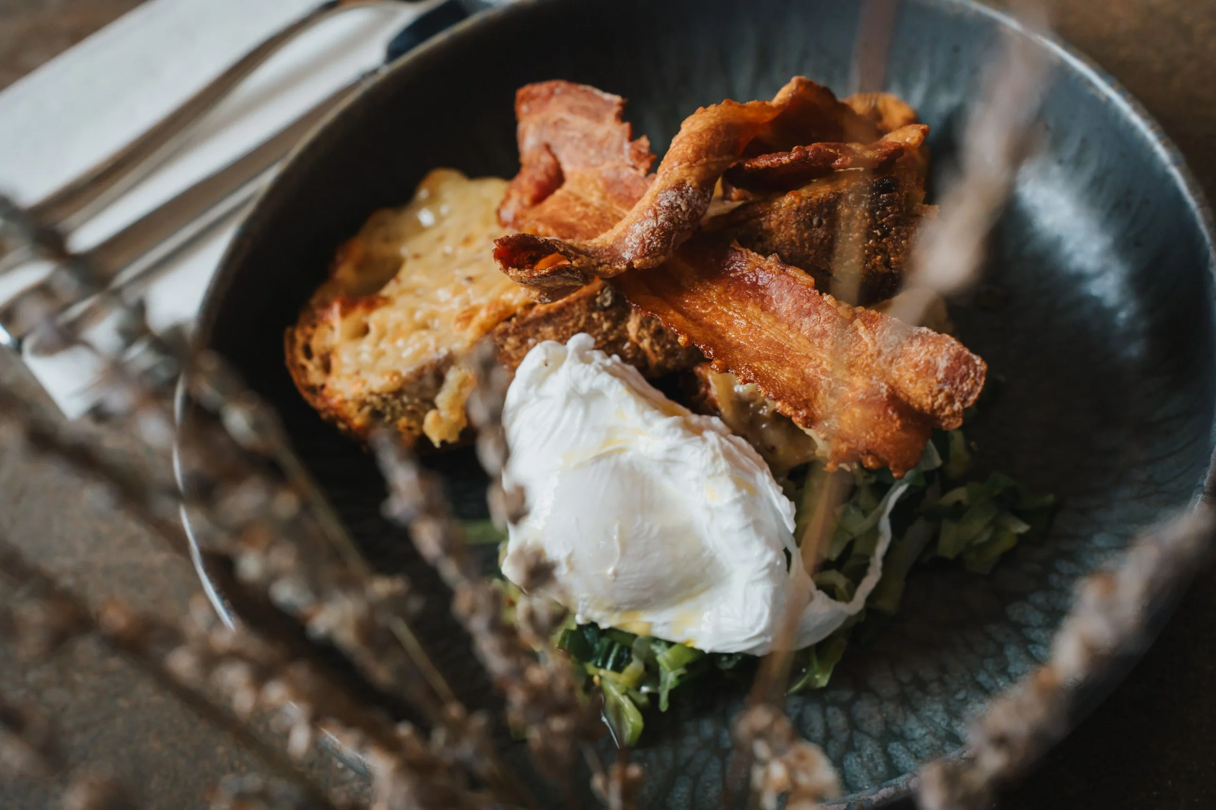 Baked macaroni and cheese with bacon, poached egg, and greens served in a dark bowl.
