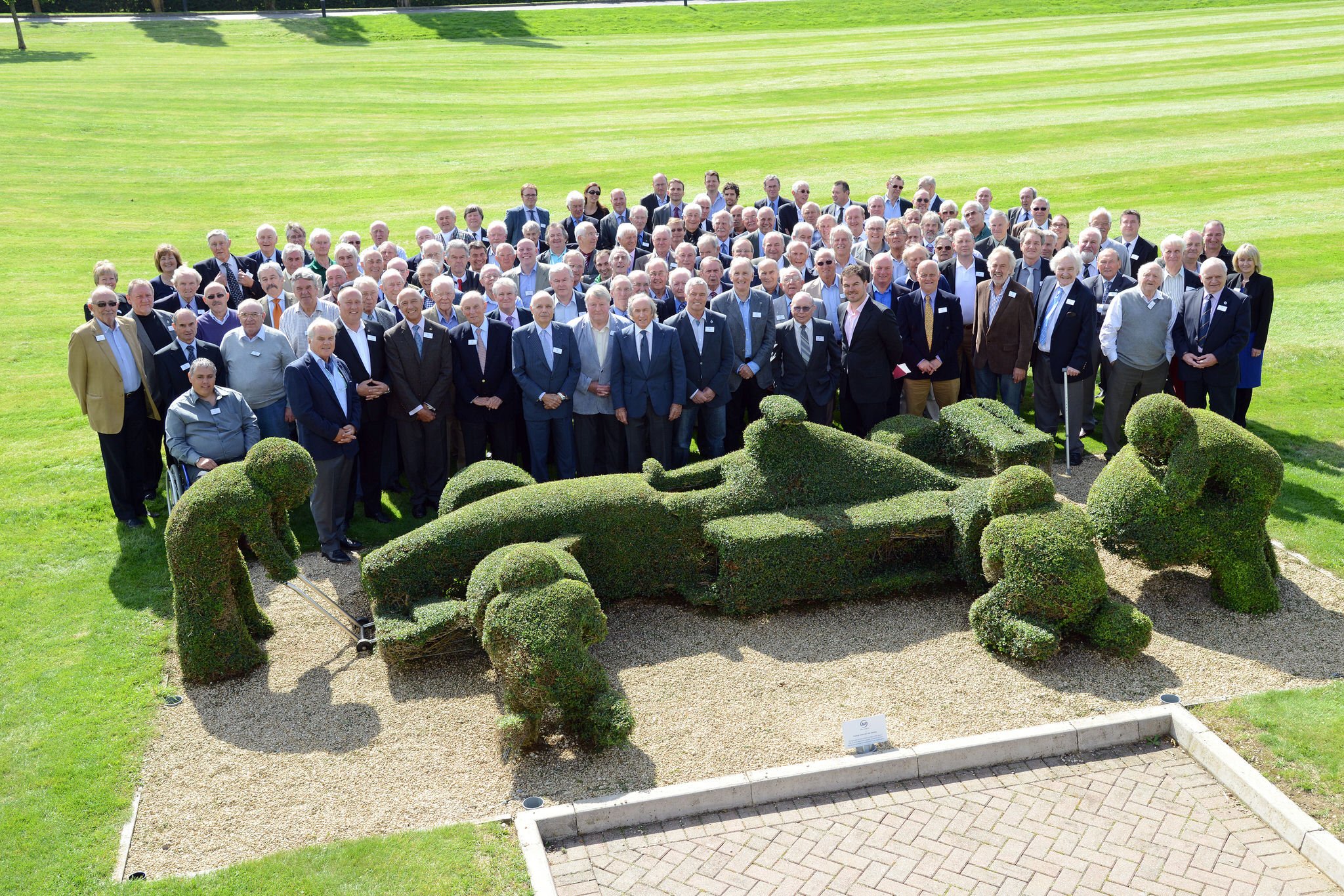 Group of people gathered outdoors in front of a large topiary sculpture resembling a race car with four dogs, on a neatly maintained lawn.