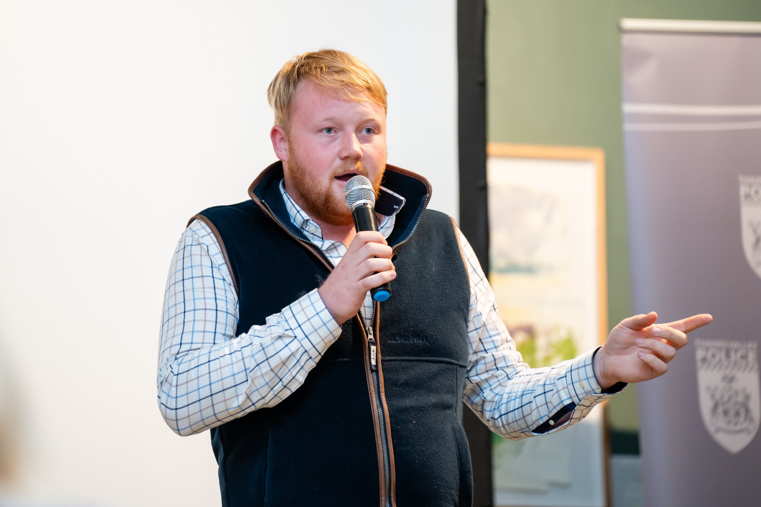 Kaleb Cooper from Clarkson's Farm speaking at a farmer fourm, wearing a checkered shirt and a black vest, pointing with his right hand.