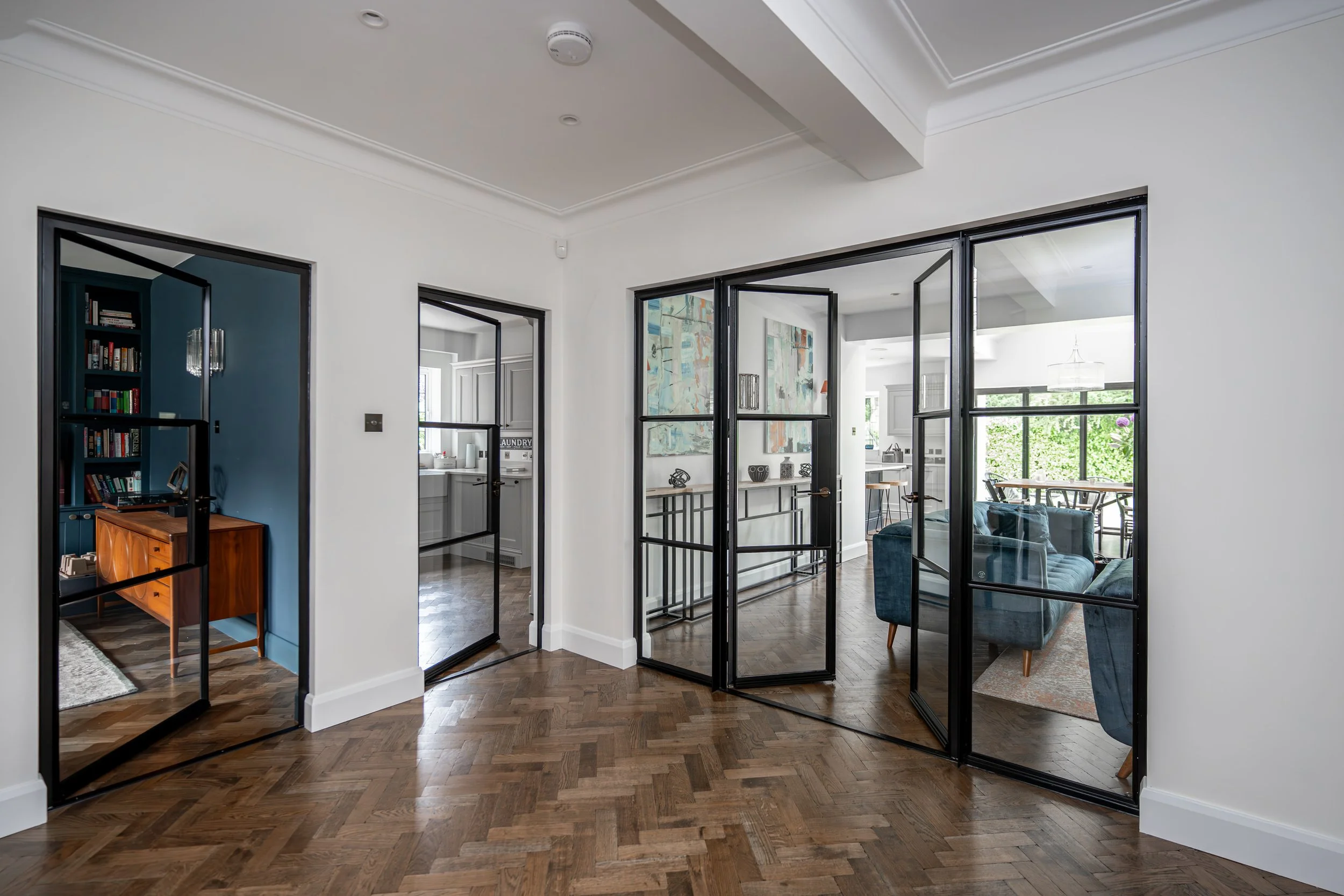 Interior view of a modern home with wood flooring, white walls, glass Crittall partition doors with black frames dividing the spaces, and visible rooms including a living area with a blue sofa, bookshelf, dining area, and kitchen.