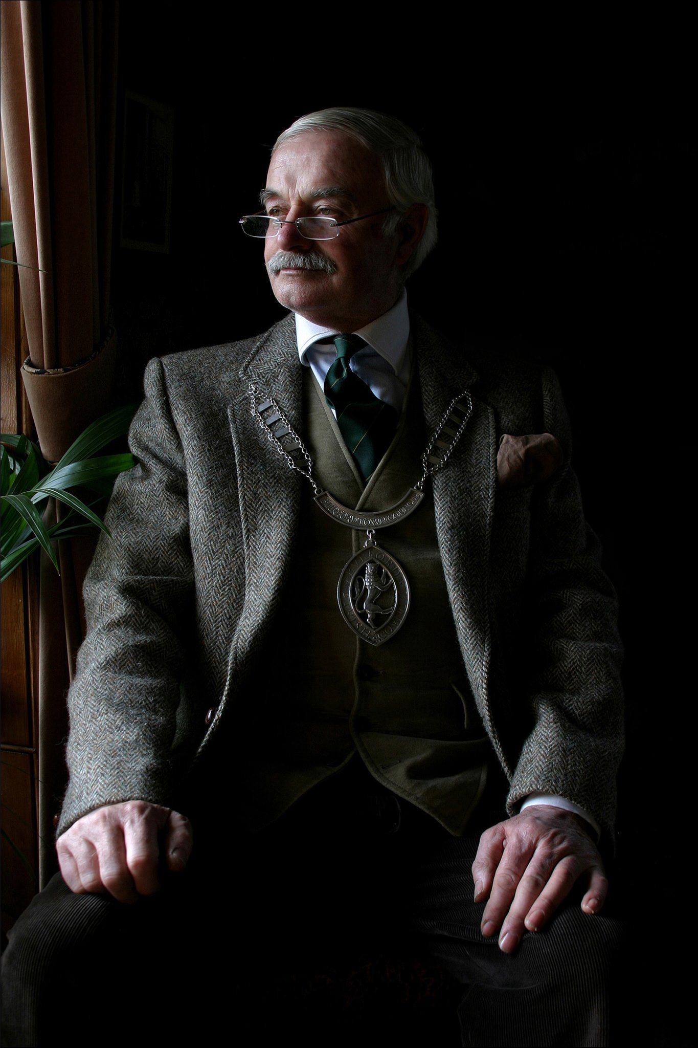 An elderly man with white hair and a mustache, wearing glasses, a brown tweed jacket, a white shirt, a dark green tie, and a patterned vest, sits by a window with brown curtains, in a dimly lit room.