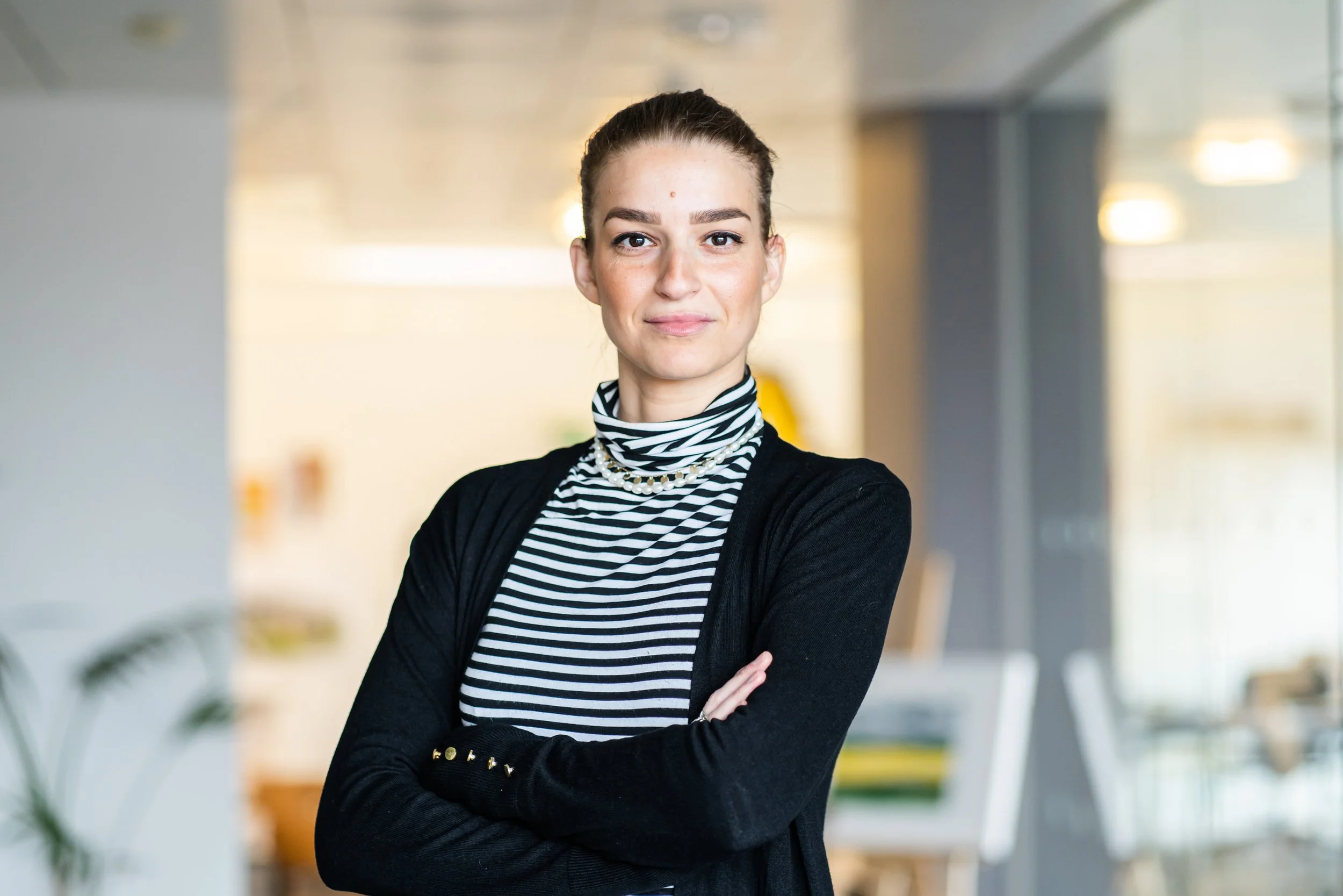 A confident woman with crossed arms in an office setting, wearing a striped turtleneck, pearl necklace, and black blazer.