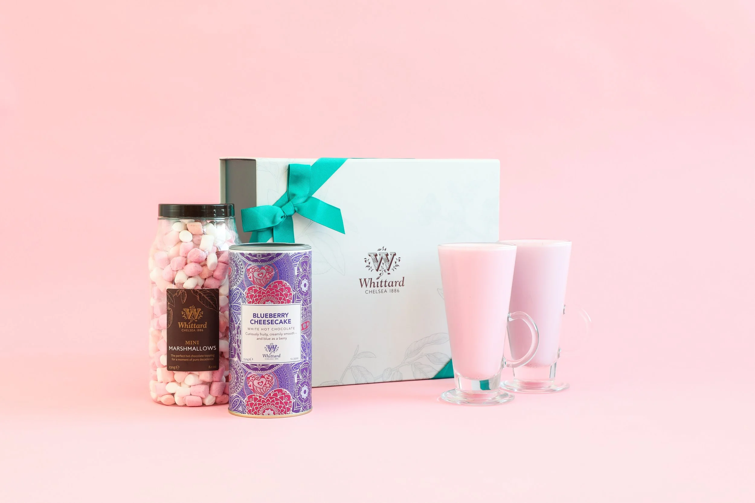 A gift box with a teal ribbon, two pink mugs, a jar of pink and white marshmallows, a can of blueberry cheesecake white chocolate, on a pink background.