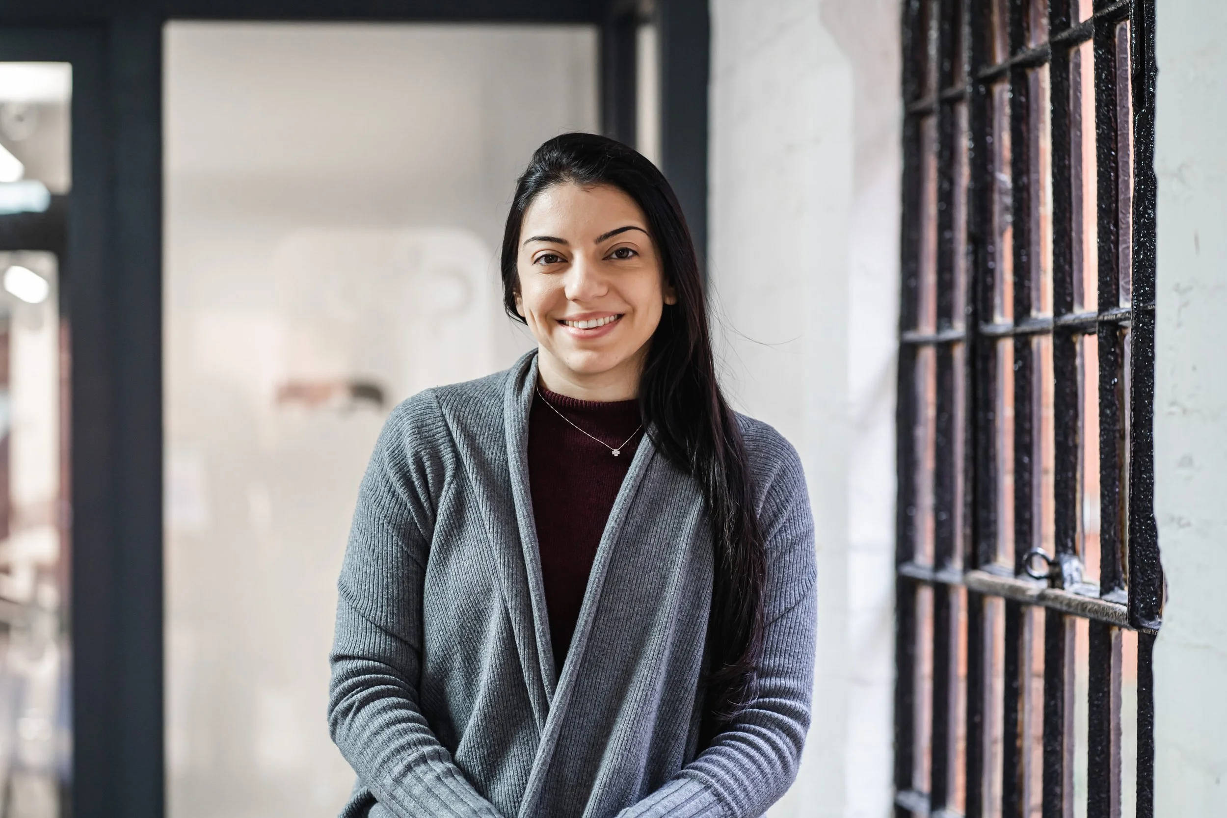 A woman with long dark hair smiling whilst posing for her company headshot profile image, standing indoors near a brick window with black iron bars, wearing a gray cardigan over a maroon top Oxford Photography
