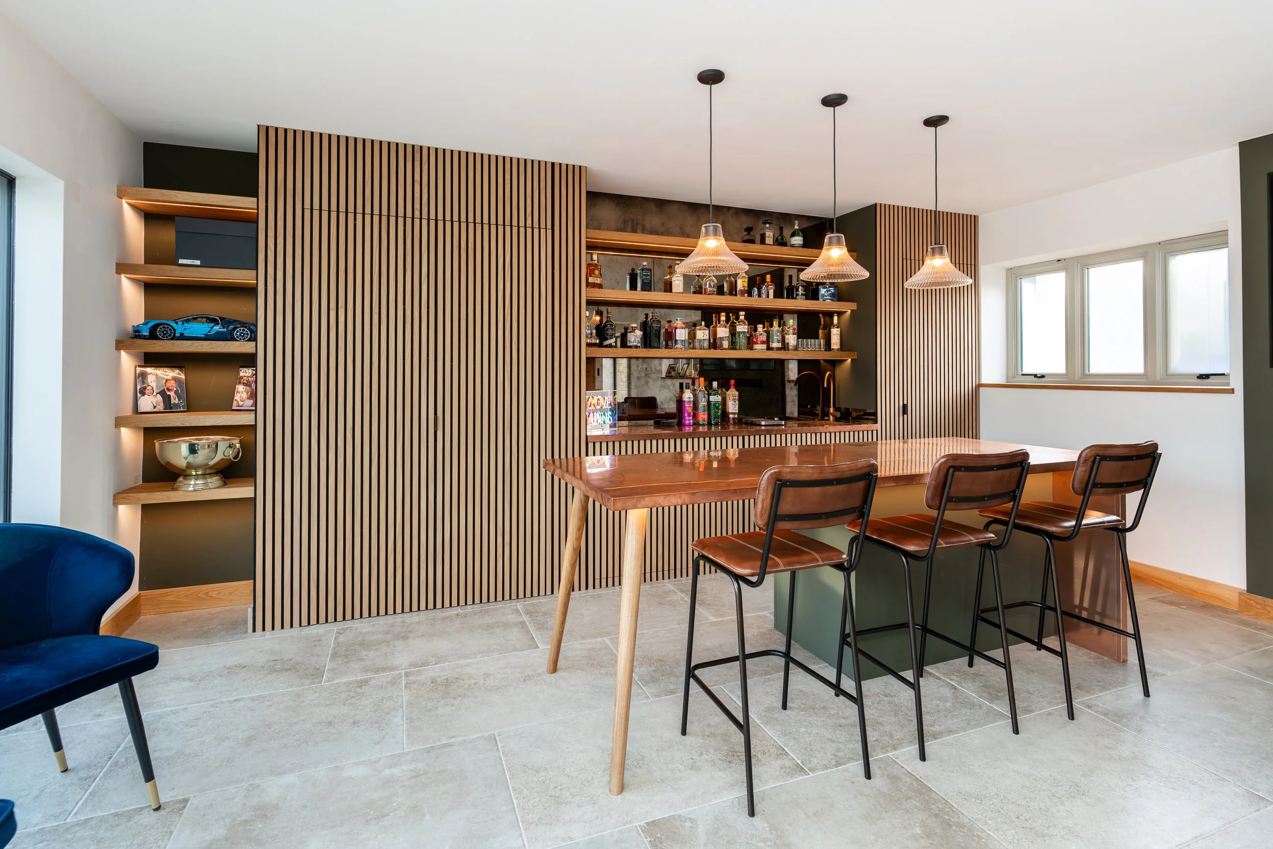 Modern kitchen with a bar, three leather chairs, a wooden table, and a built-in wooden shelving unit with liquor bottles and decorative items.