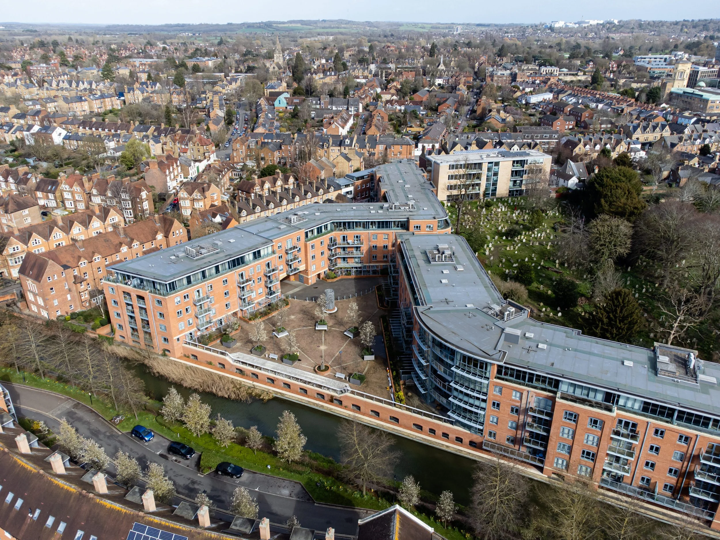 Aerial view of a modern apartment complex beside a small waterway in a residential neighborhood with houses and trees in the background.