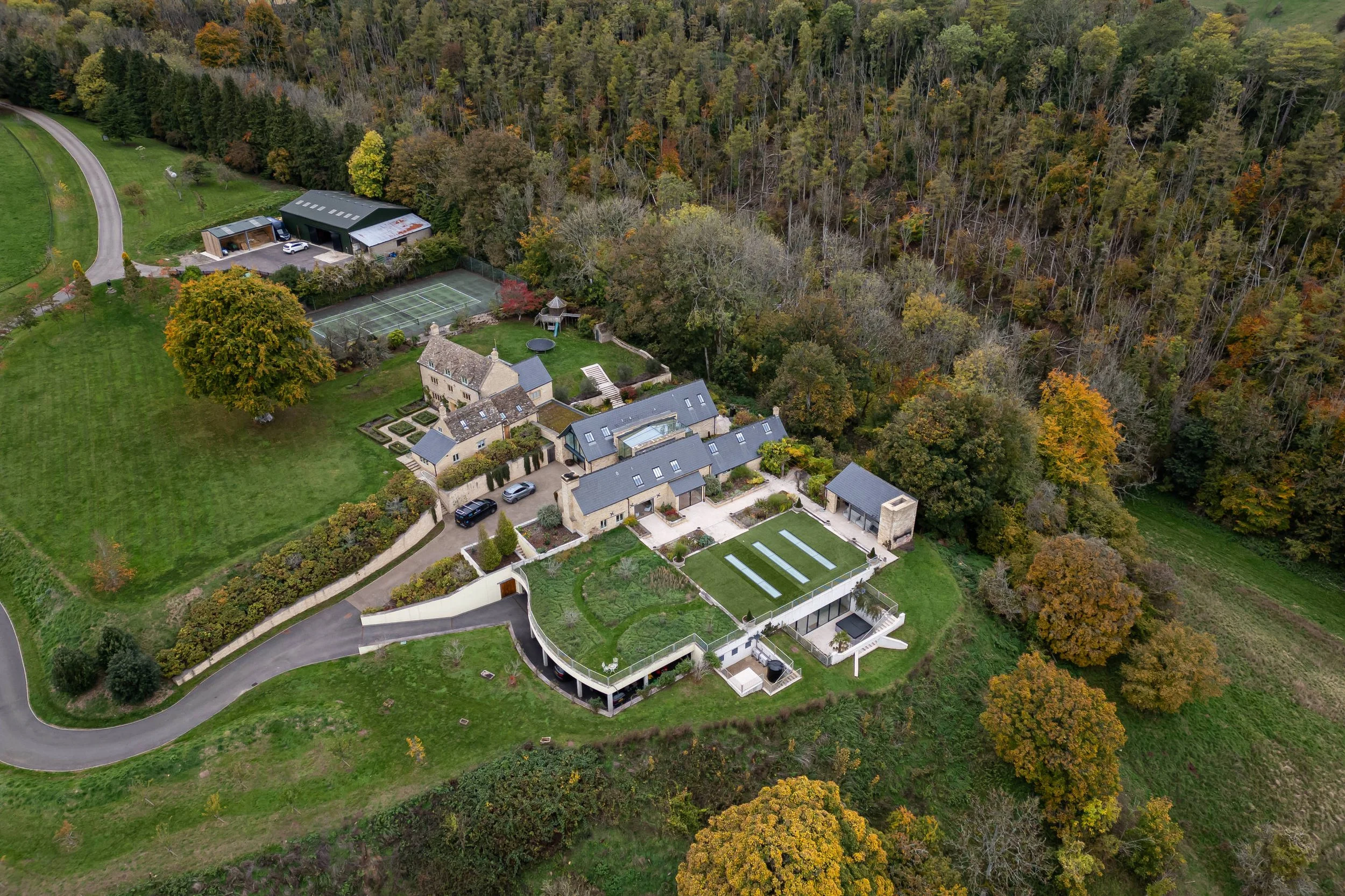 Aerial view of a large estate with multiple buildings, manicured gardens, a parking area with cars, a tennis court, a trampoline, and surrounding trees with fall foliage.