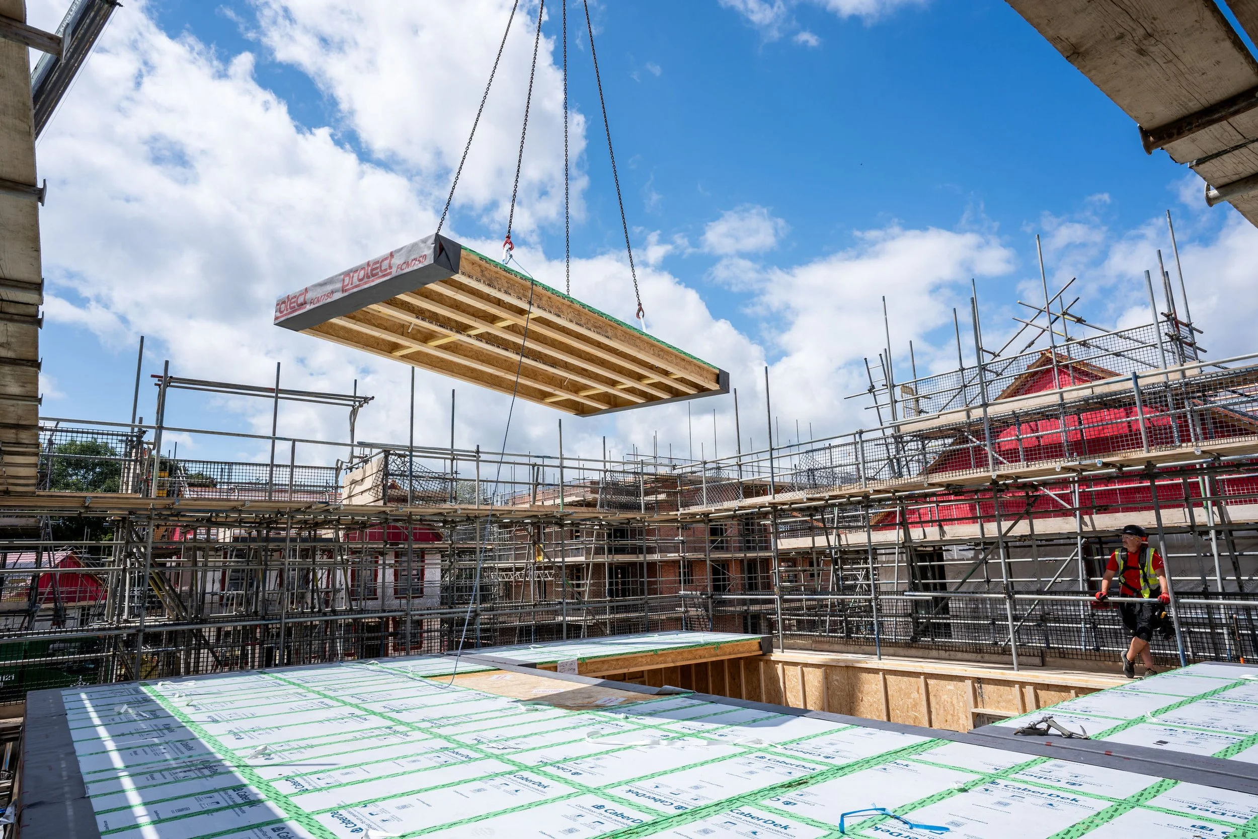 Construction site with scaffolding, with a large wooden panel being hoisted by a crane against a blue sky with clouds.