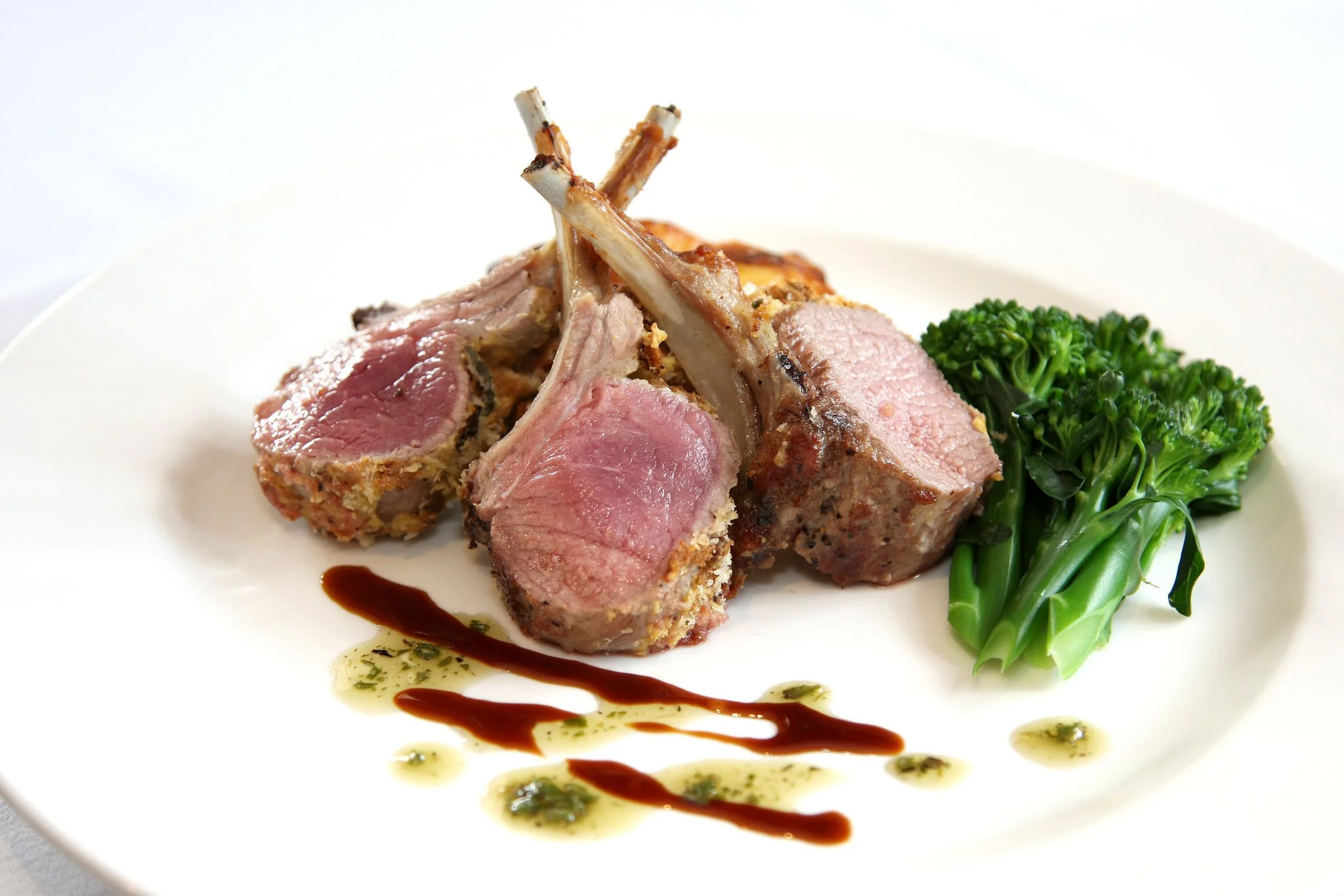 A plate of cooked rack of lamb with pink interior, garnished with green broccoli and drizzled with brown sauce and green herb oil.