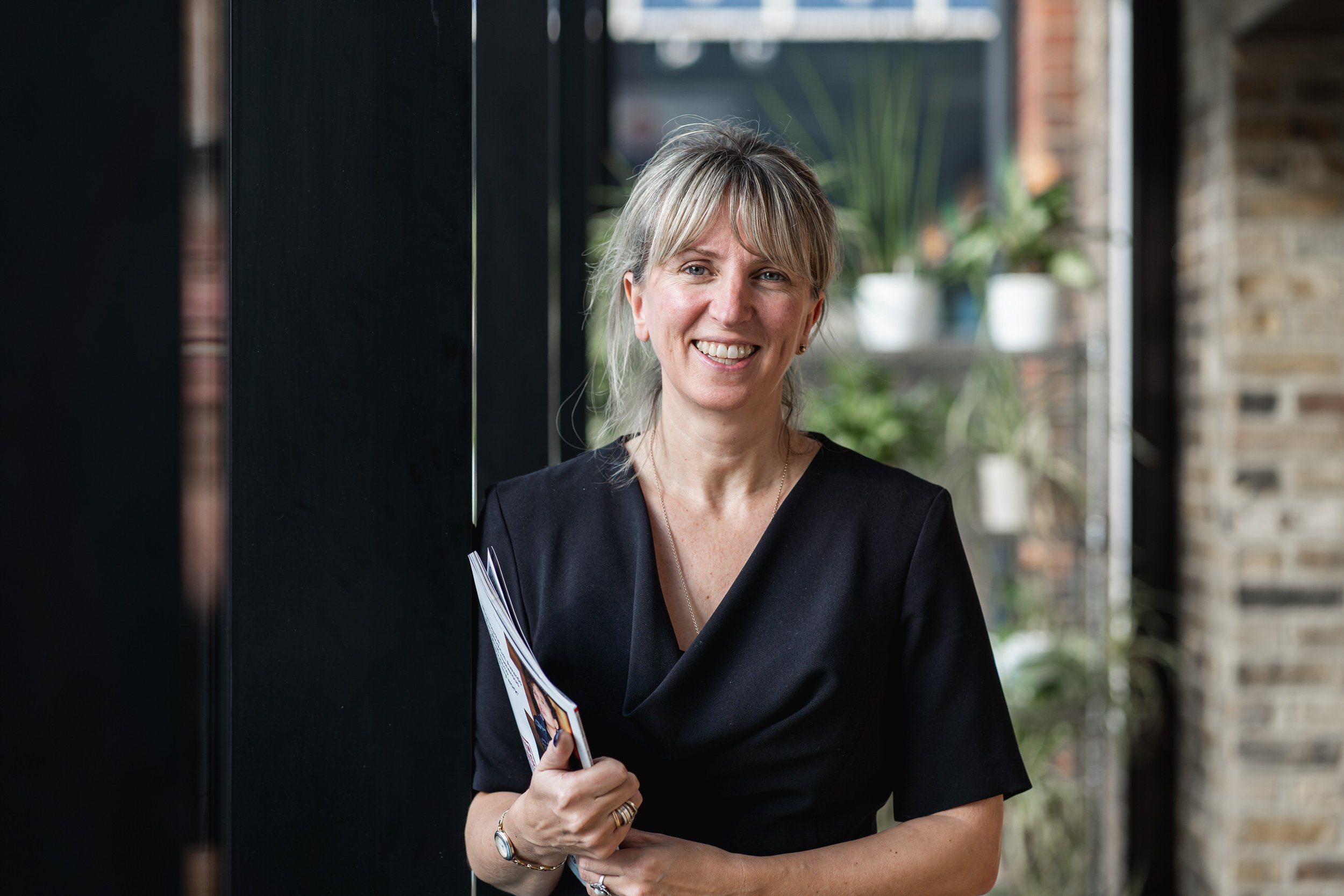 A smiling woman with blonde hair and a black dress standing indoors, holding a magazine or brochure, with a window and plant in the background.