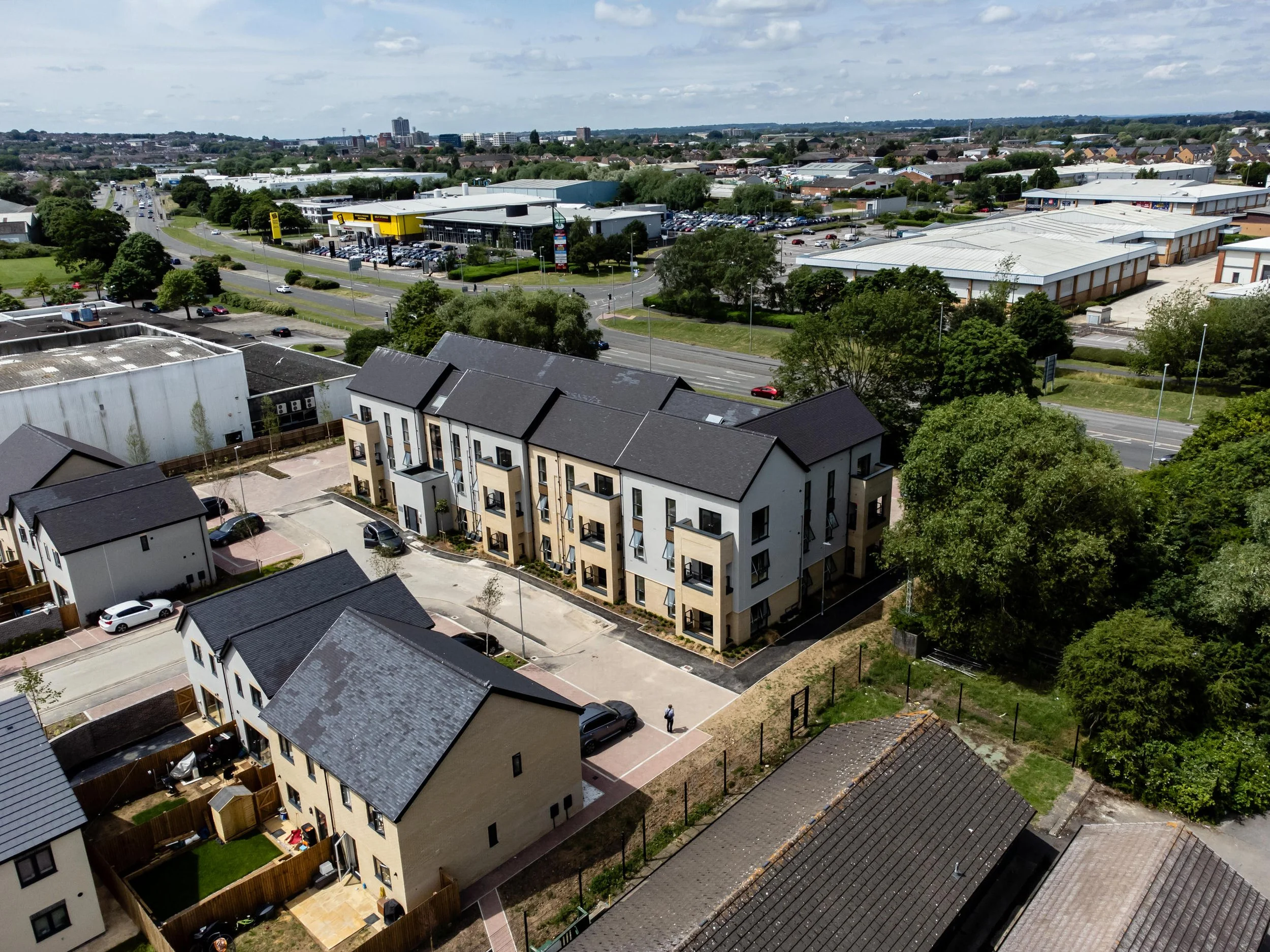 An aerial view of residential apartment buildings with parking spaces, surrounded by trees, and commercial area with warehouses and retail stores in the background.