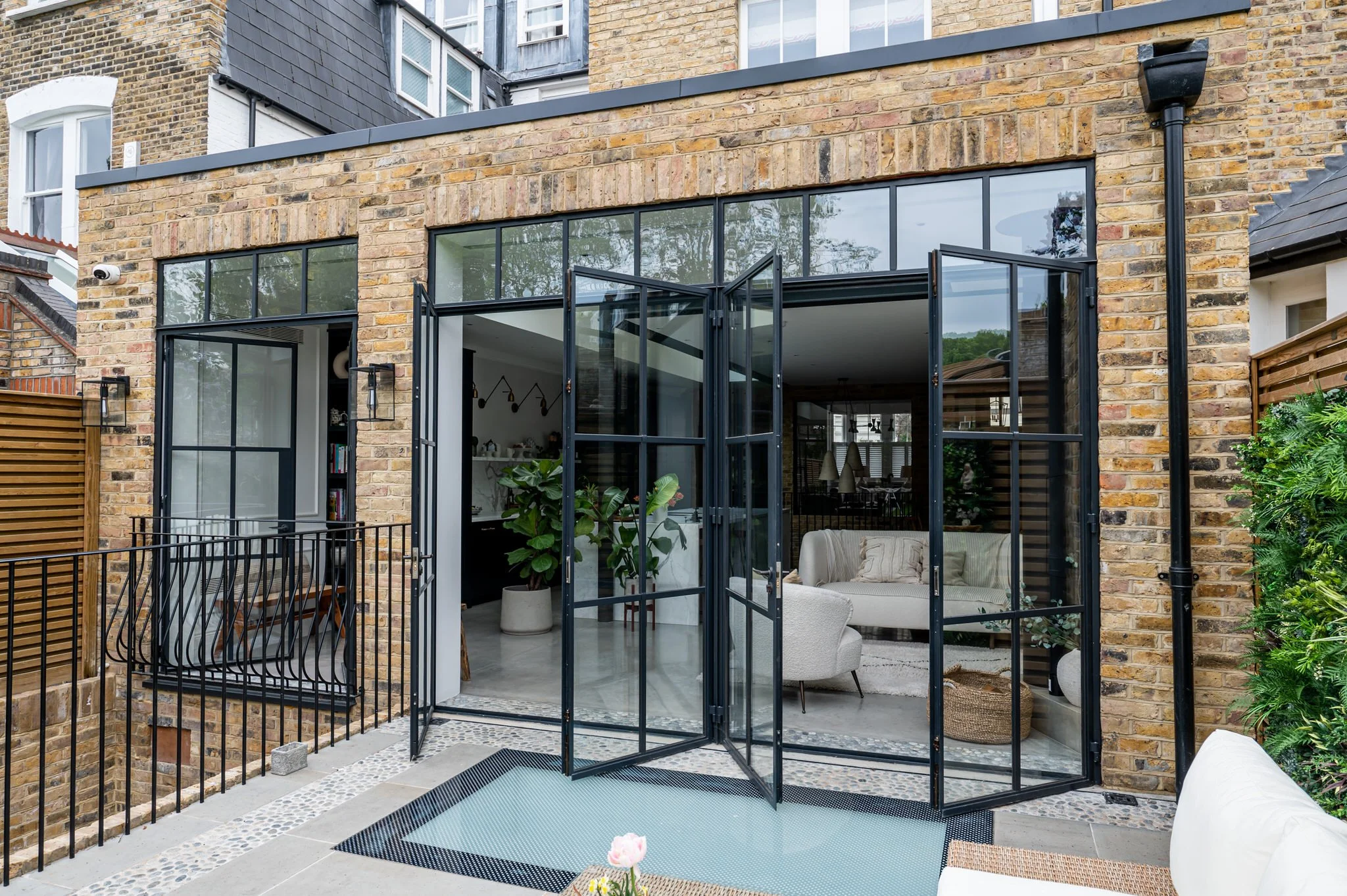 Modern brick house with large black-framed Crittall glass doors opening to a patio with outdoor seating and greenery.
