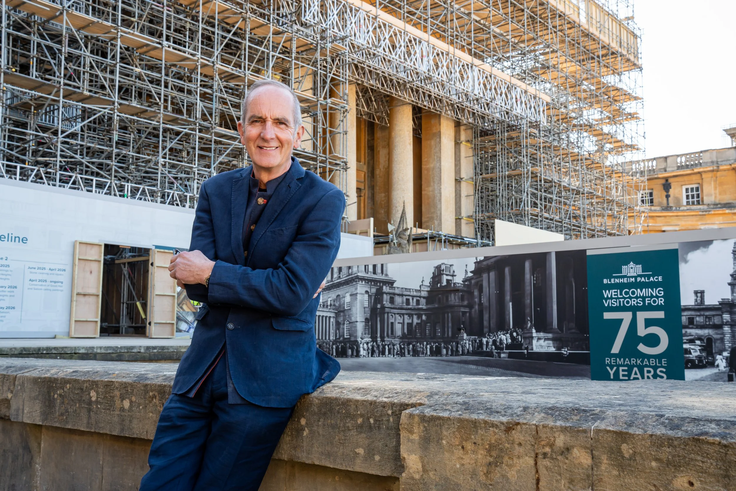 A man in a navy blue suit leaning against a stone wall in front of a building under renovation with scaffolding, and a sign mentioning 75 years of visitors at Blenheim Palace.