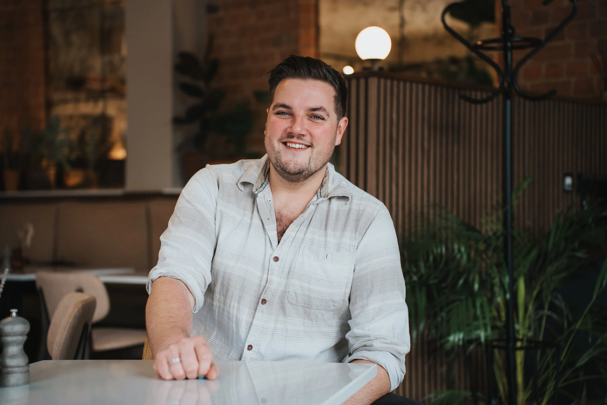 A smiling man in a white button-up shirt sitting at a table in a cozy, modern restaurant or cafe with warm lighting and wooden decor.