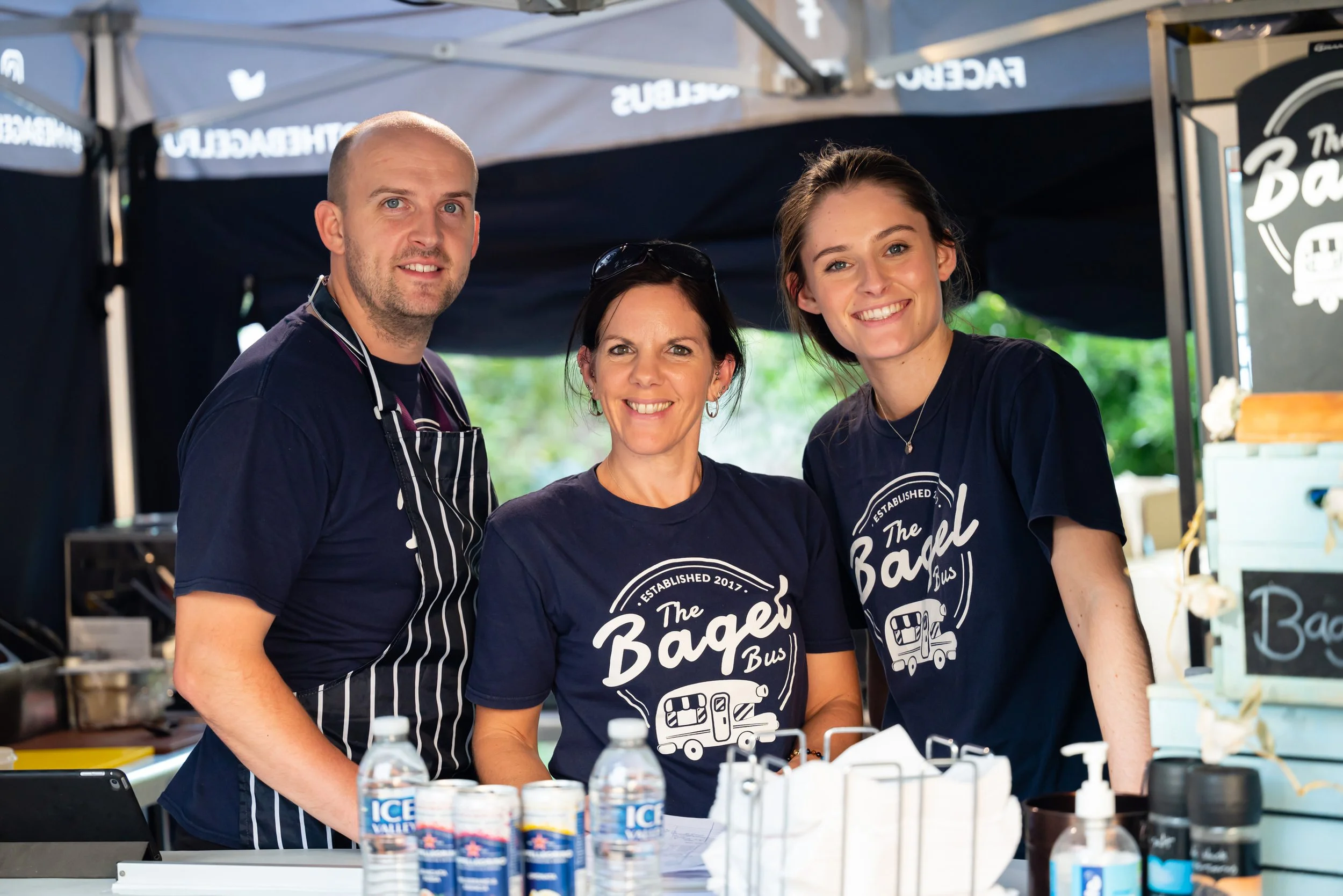Three smiling people working at a food stall named 'The Bagel Bus' wearing navy blue t-shirts with the cafe's logo, inside a black tent at a street fair or market.