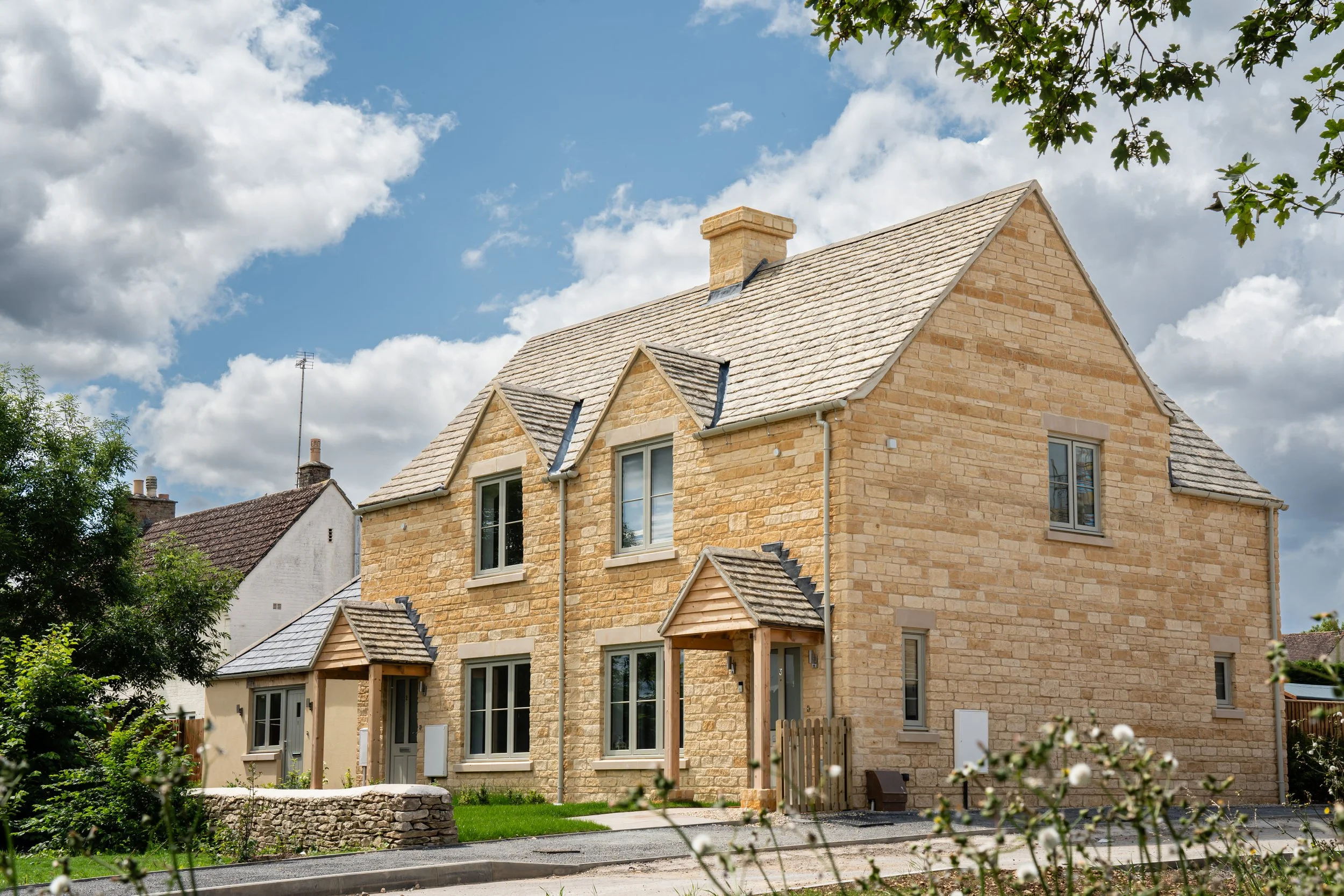 A two-story stone house with a pitched roof, small front porch, and multiple windows. The house is situated in a suburban neighborhood with greenery and a cloudy blue sky overhead.