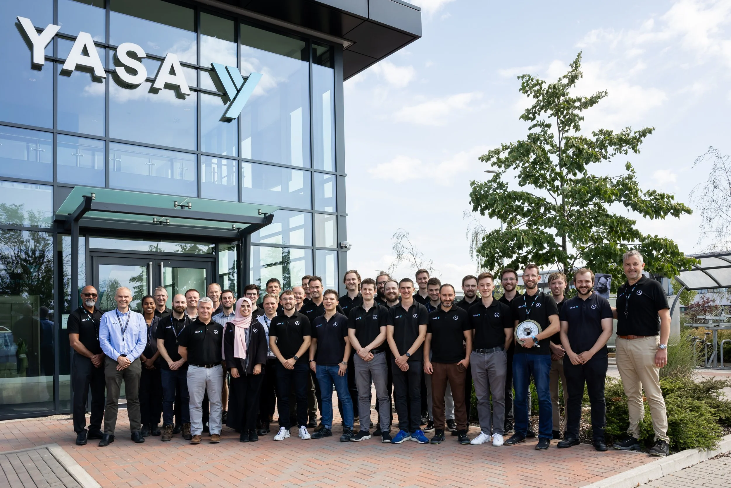 Group of people standing outside a modern glass building with 'YASA' sign, smiling, in a sunny day with greenery.