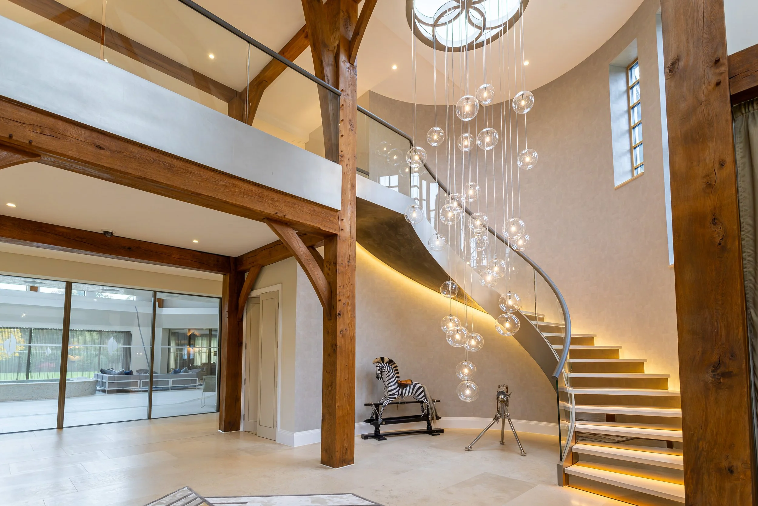 Interior of a modern house featuring a wooden staircase with hanging globe pendant lights, a curved wall, and a mezzanine with a glass railing. There is a zebra rocking horse and a tripod telescope on the lower level.