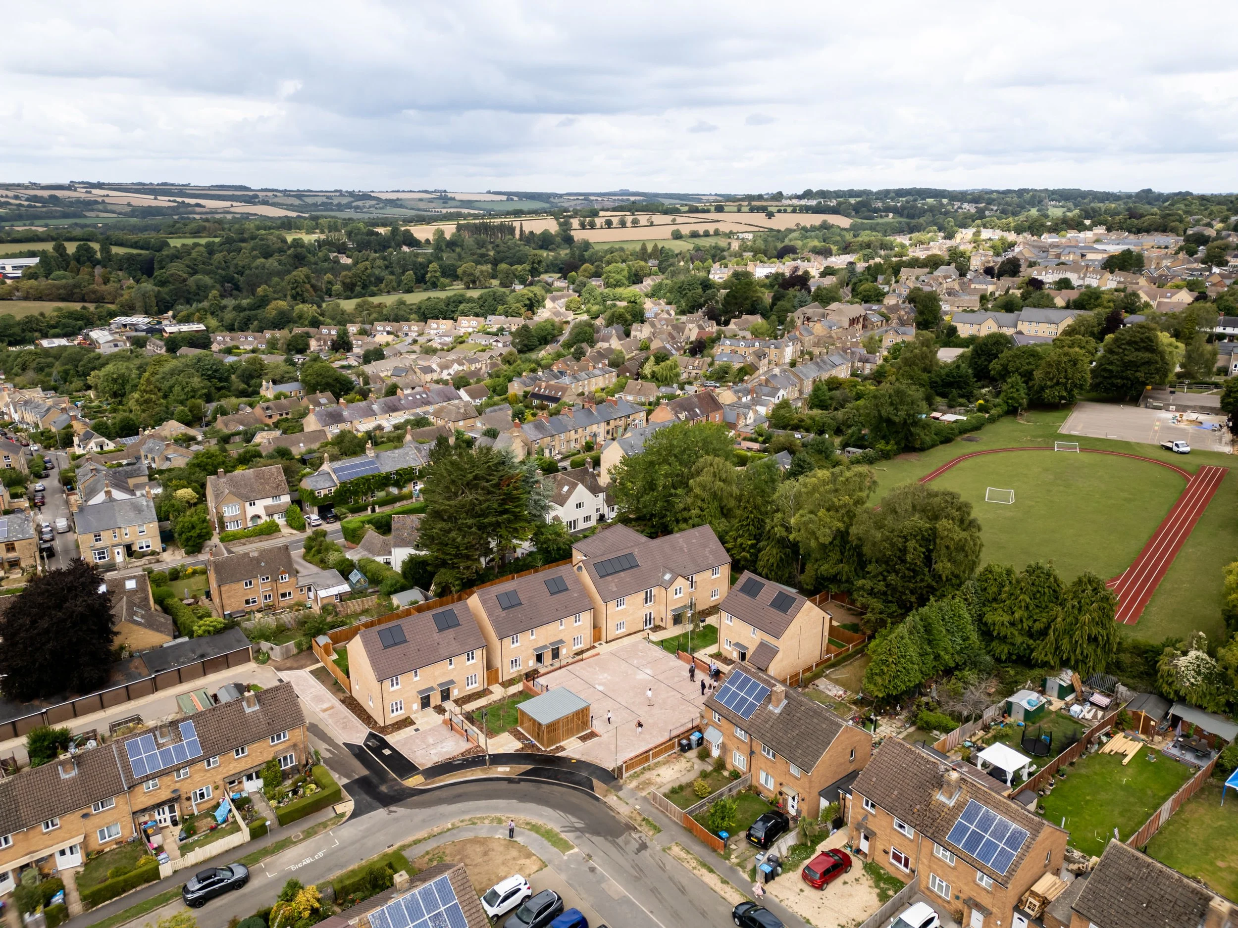 Aerial view of a suburban neighborhood with a schoolyard, sports field, houses, and green spaces under a cloudy sky.