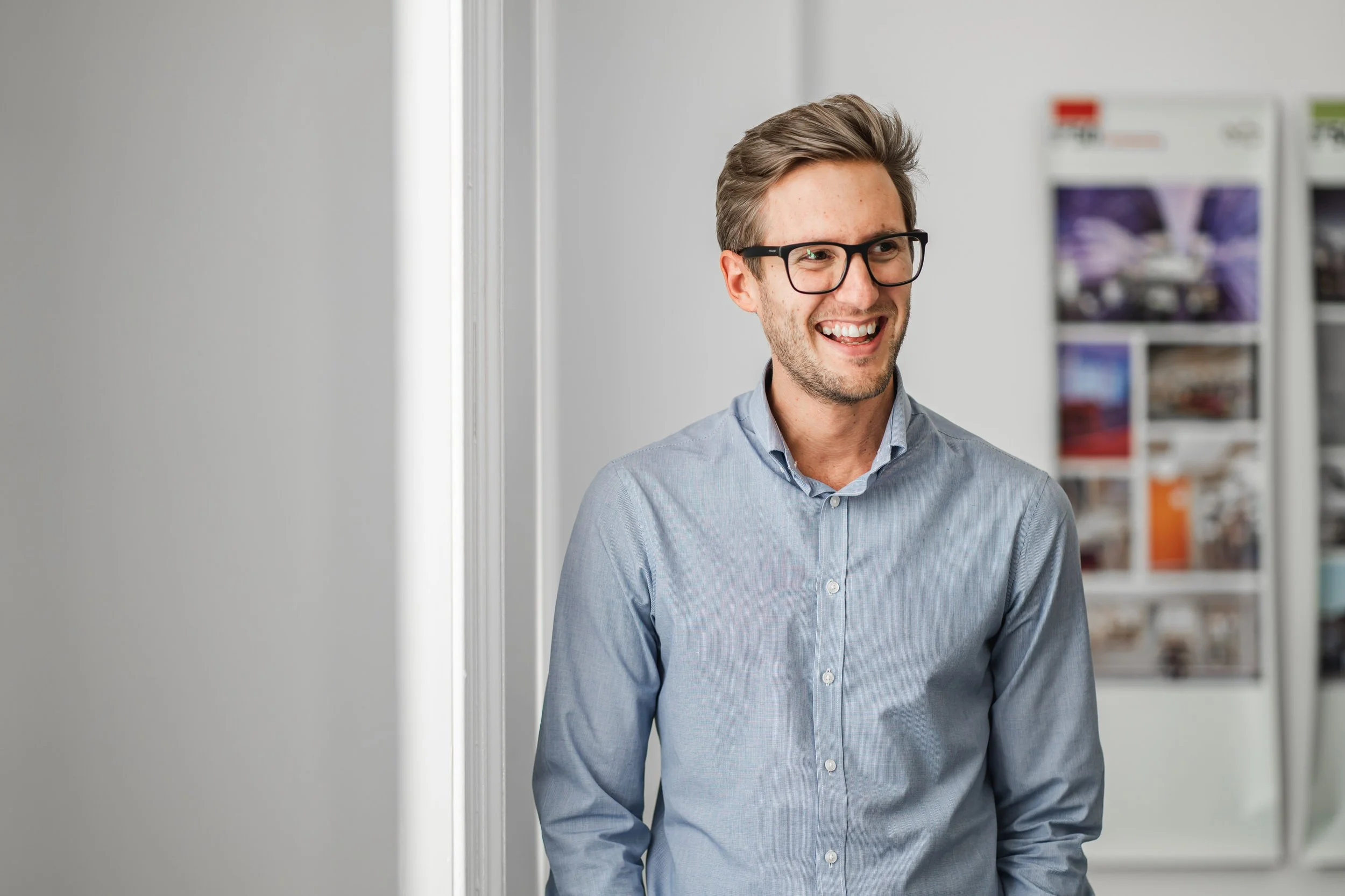 A smiling man having his LinedIn team picture taken with glasses and a light blue button-up shirt standing indoors near a wall with a bulletin board or magazine rack in the background.