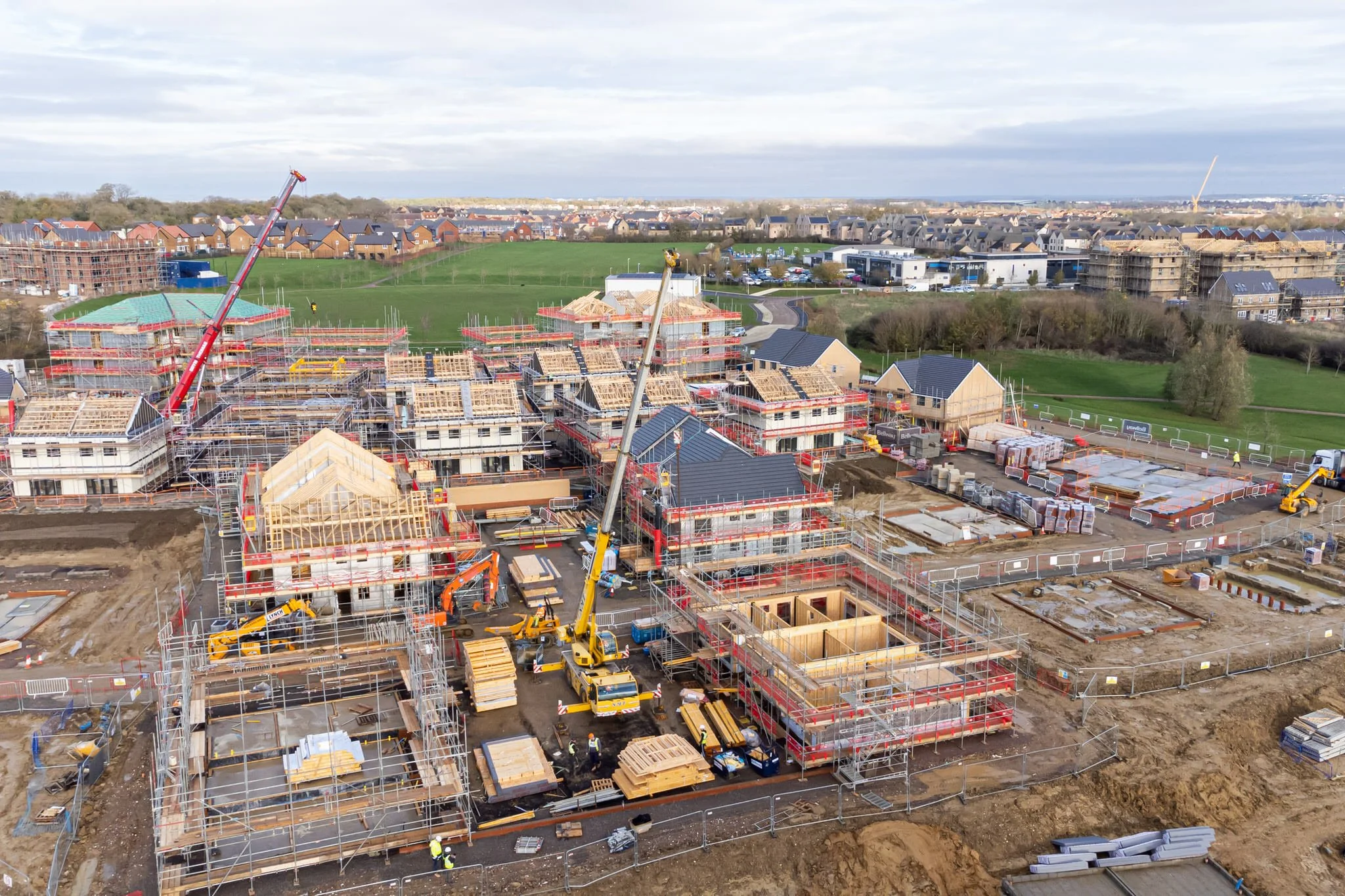 Construction site with multiple buildings under construction, cranes, scaffolding, construction workers, and building materials. In the background, there are completed houses and green fields.