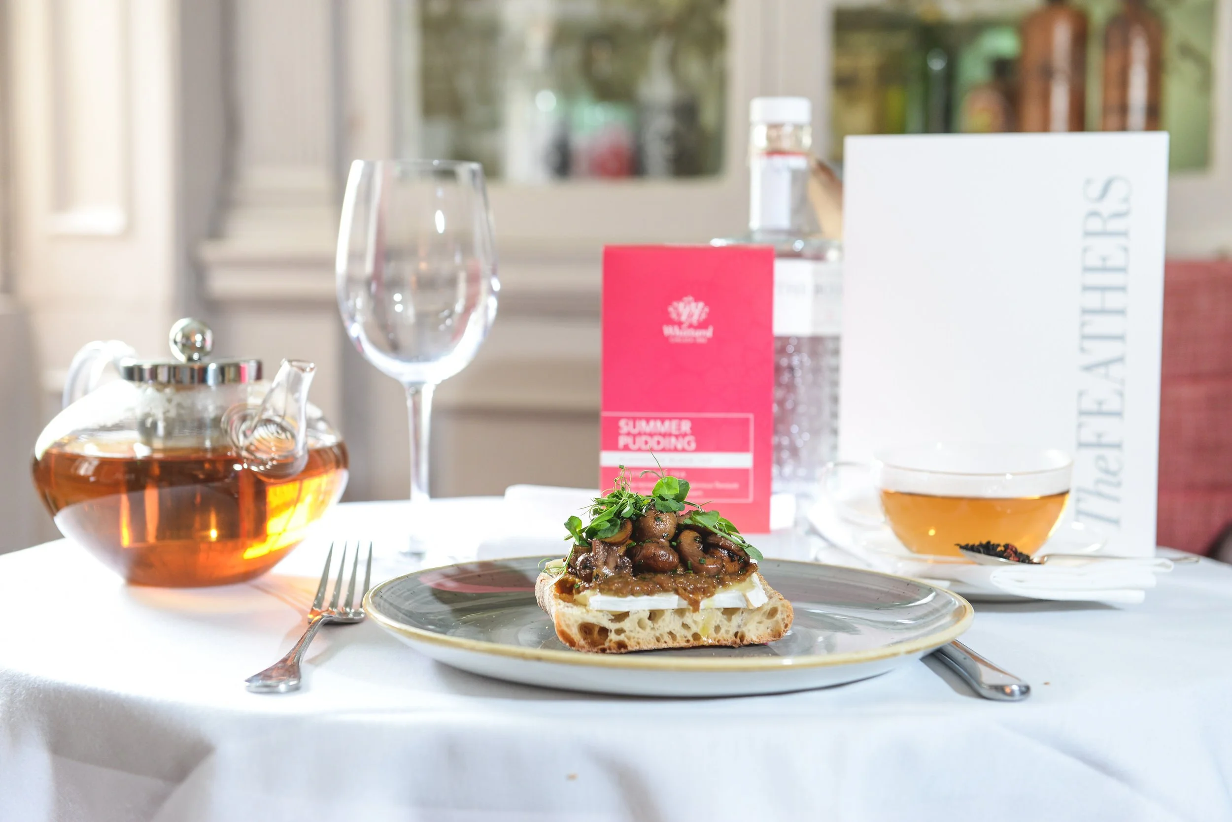 A table setting with a piece of bread topped with cream cheese, mushroom pâté, and microgreens, a teapot filled with tea, a wine glass, a bottle of water, a trolly of summer pudding, a cup of tea, and a white menu or booklet, all set on a white table