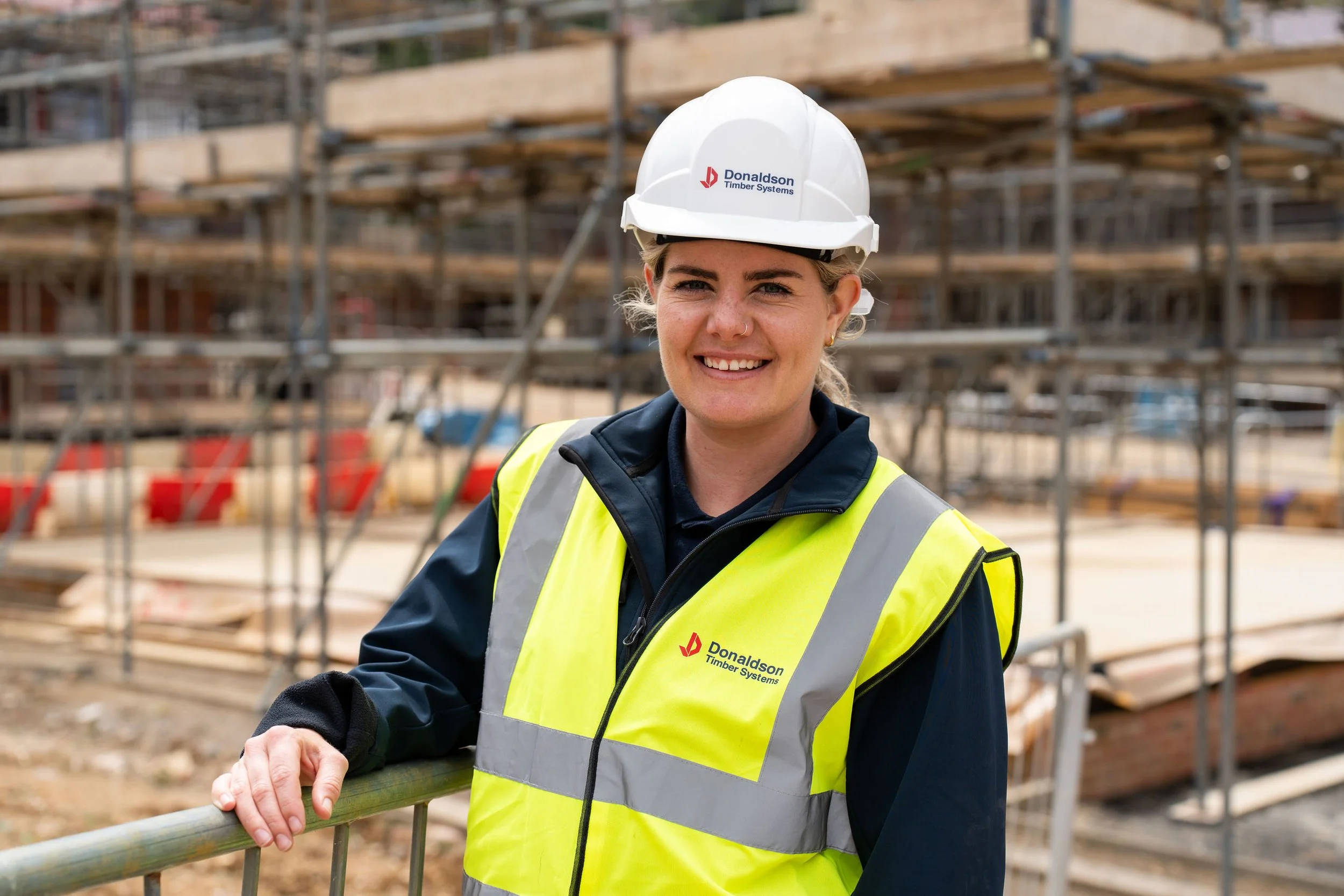A young woman construction worker wearing a white safety helmet and a yellow reflective vest with the logo 'Donaldson Timber Systems' on it, smiling at the camera at a construction site with scaffolding and lumber in the background.