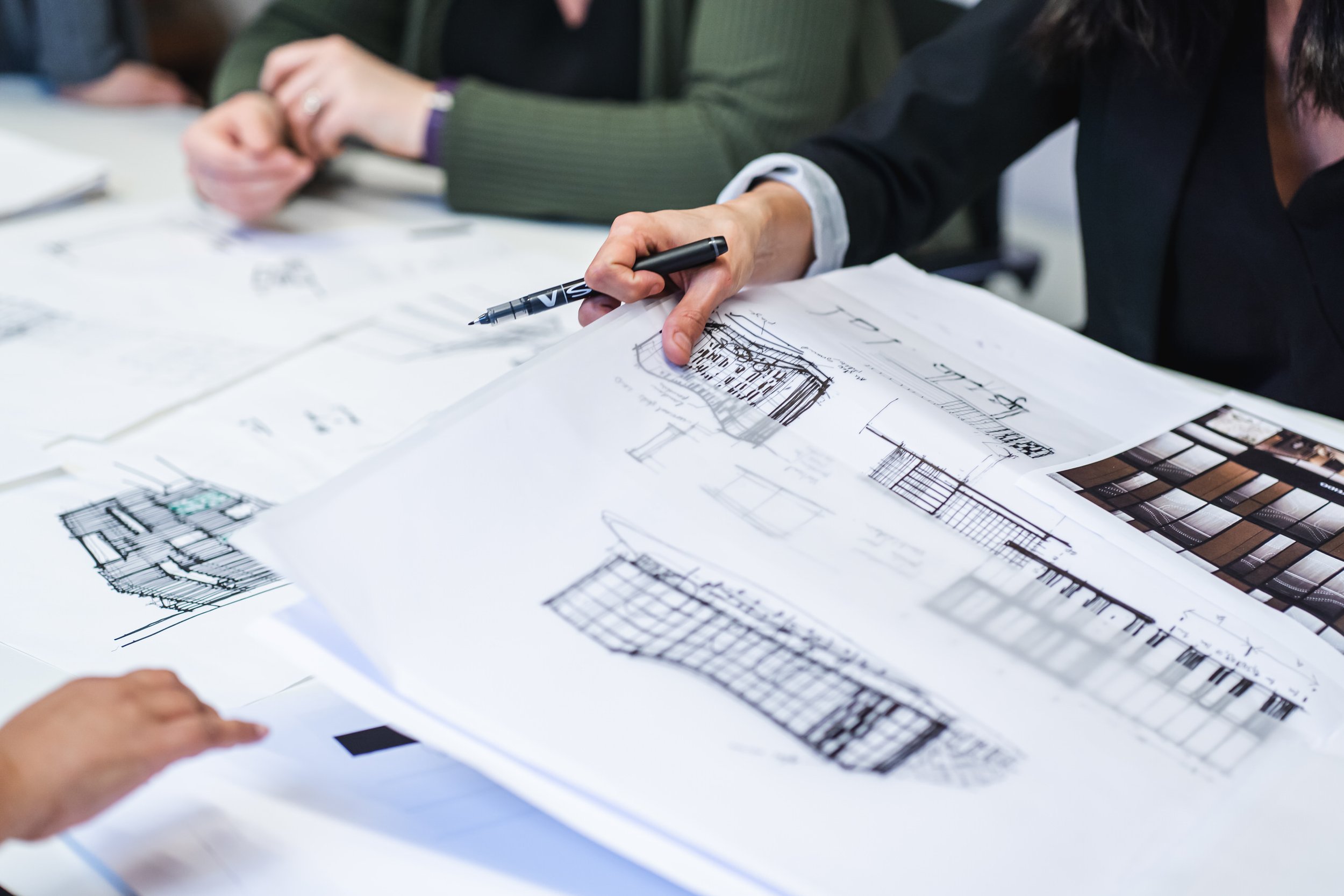 A person in a black jacket reviewing architectural plans with other individuals, examining detailed drawings and design layouts on a table.