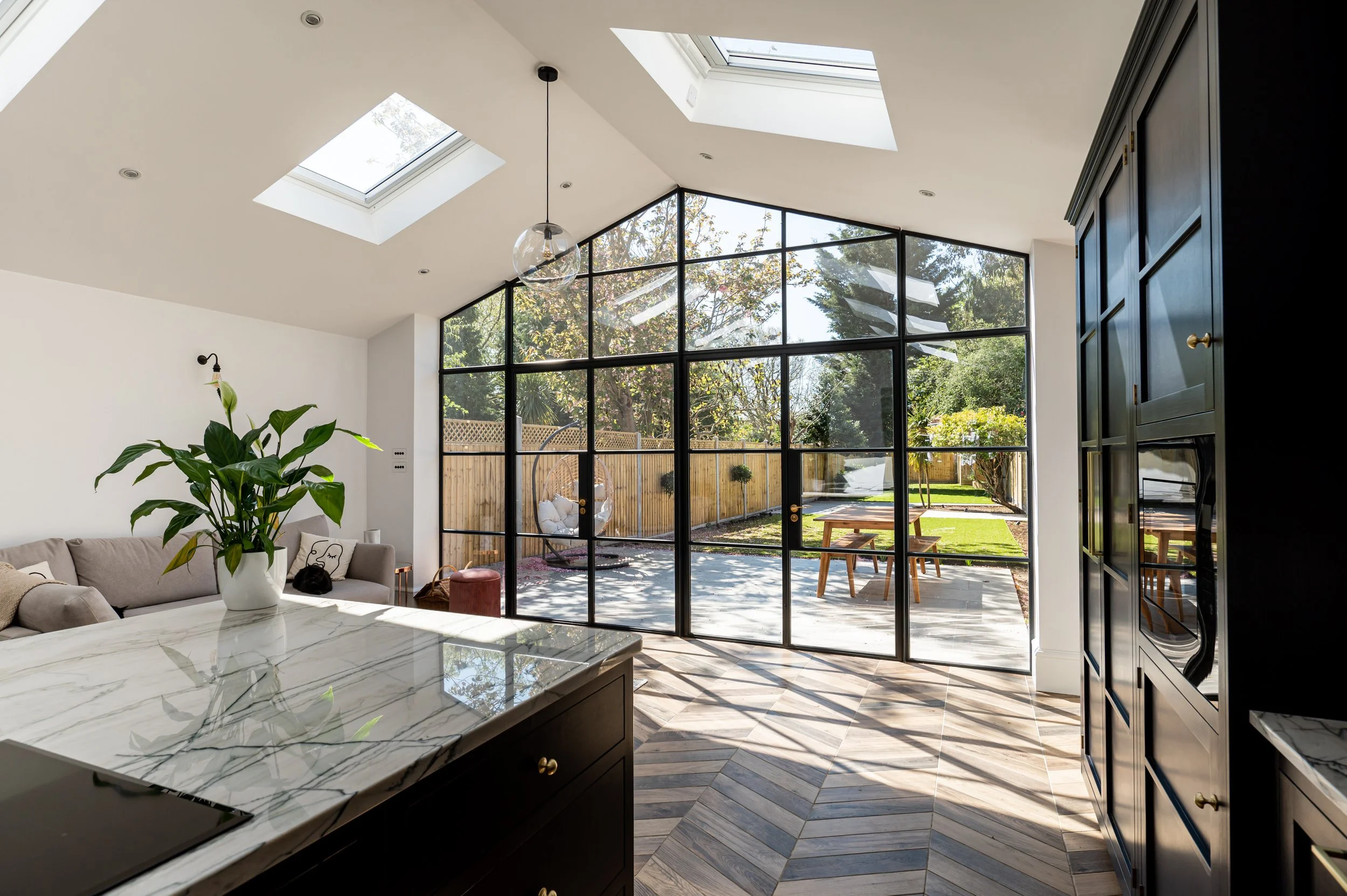 Bright modern kitchen with large black-framed glass windows, skylights, a marble countertop, and outdoor garden patio.