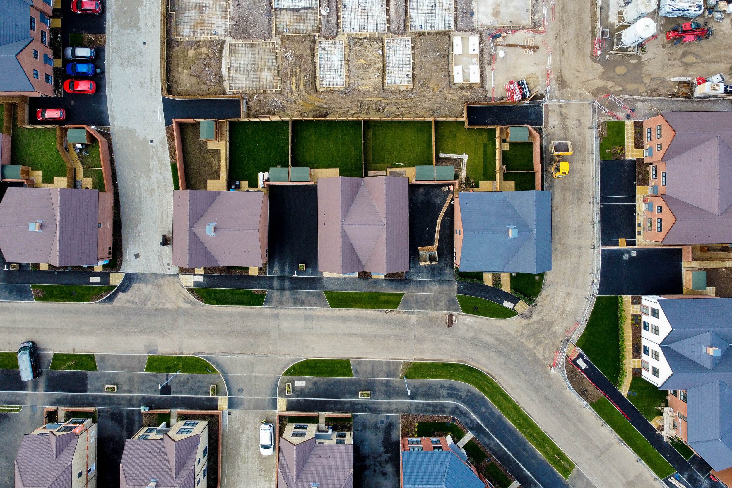 An aerial view of a residential neighborhood under construction, showing completed houses with gray and blue roofs, roads, driveways, and some construction sites with dirt and building materials.