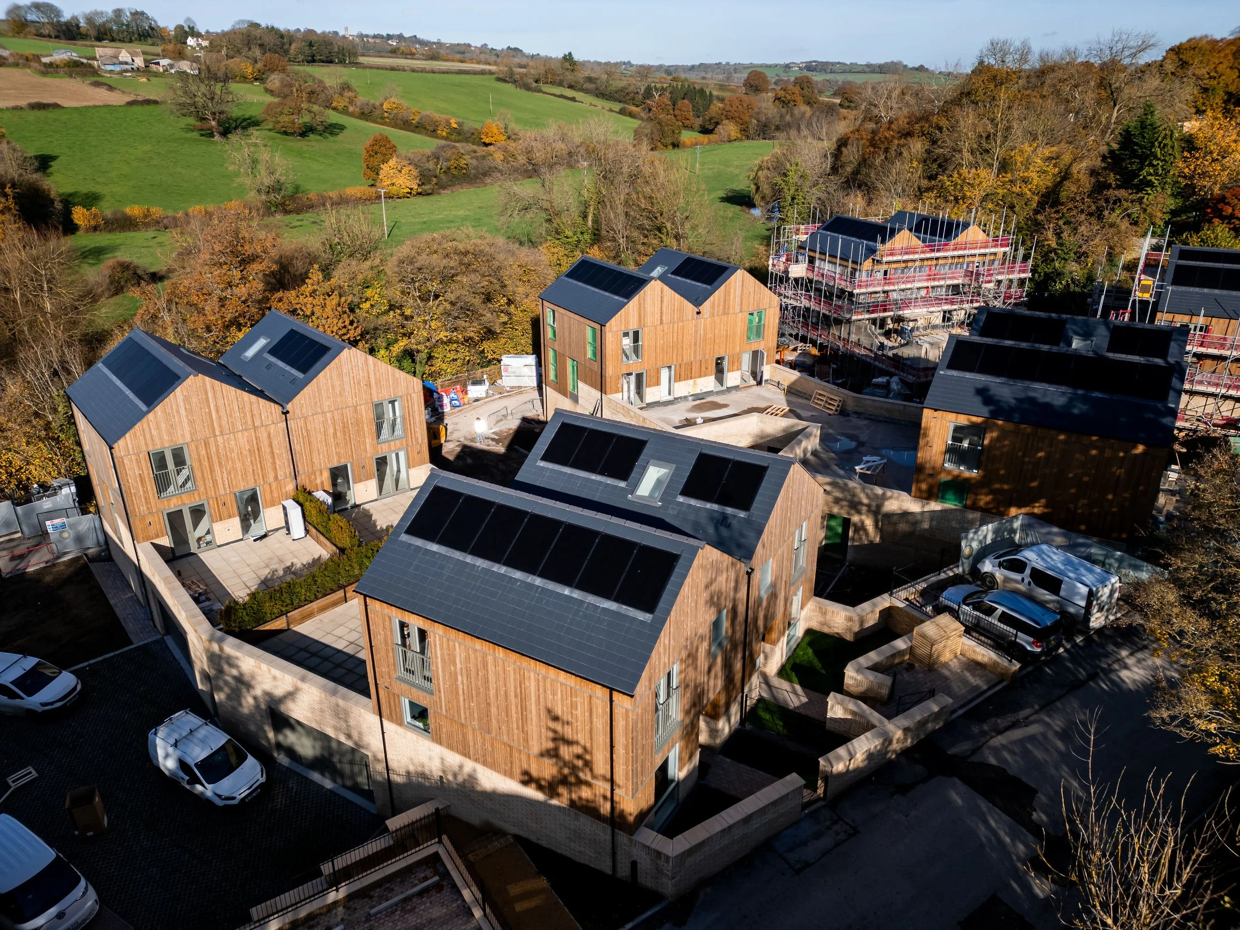 From an aerial view, modern wooden houses with solar panels on roofs in a developing residential area surrounded by trees and open green fields.