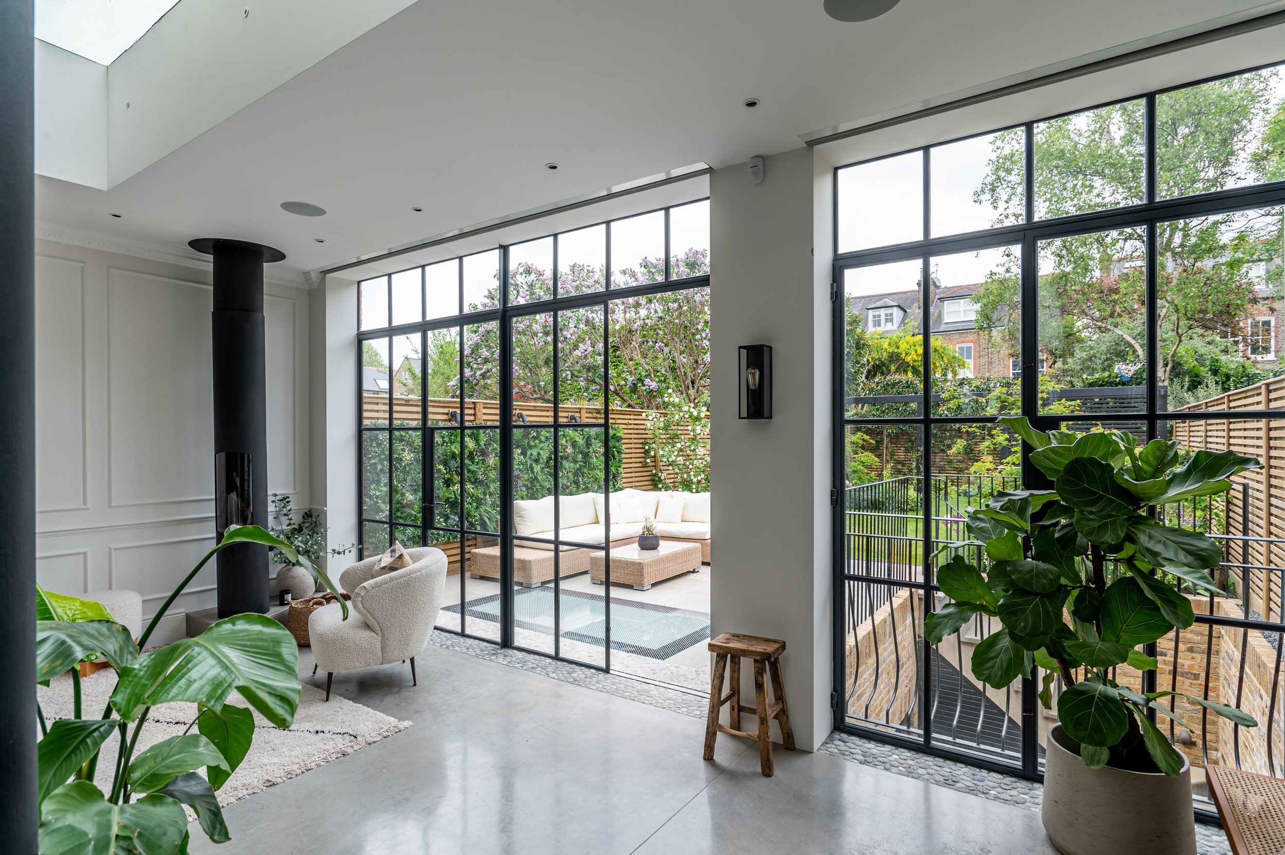 Modern living room with large glass Crittall windows and doors leading to an outdoor patio with greenery. The room features white walls, a wood stove, beige armchairs, a large potted plant, and minimal decor.