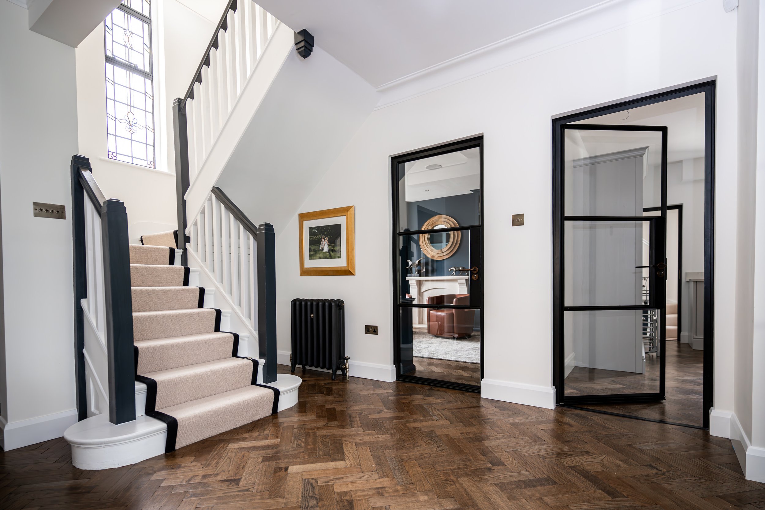 Interior of a modern house with a staircase, glass Crittall doors with black frames, framed pictures on the wall, wooden floor, and a radiator near the wall.
