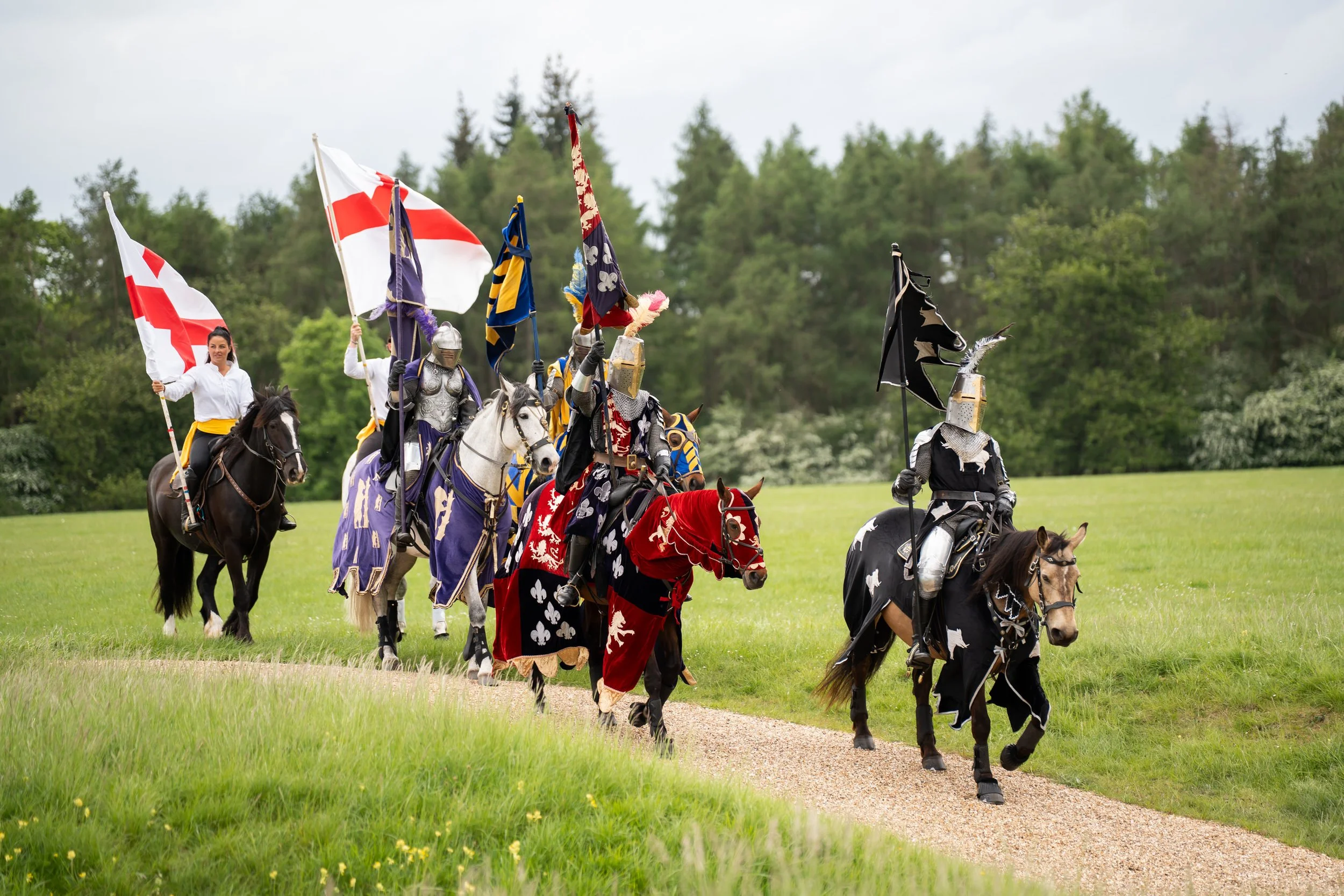 Group of armored knights and a woman on horseback holding flags, riding along a grassy path with trees in the background.