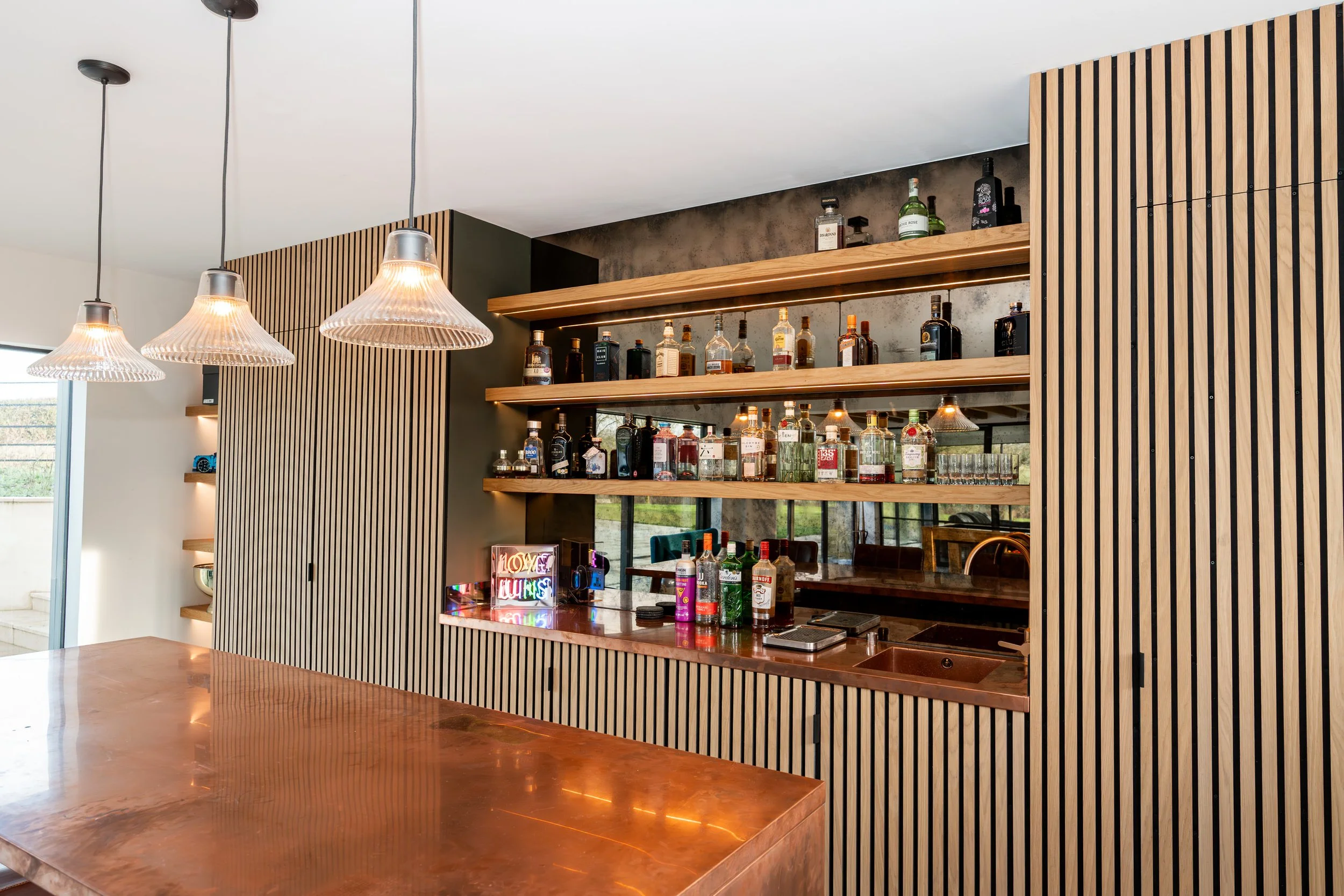 Modern bar area with wooden shelves holding various bottles of alcohol, a wooden bar counter, and modern pendant lights hanging from the ceiling.