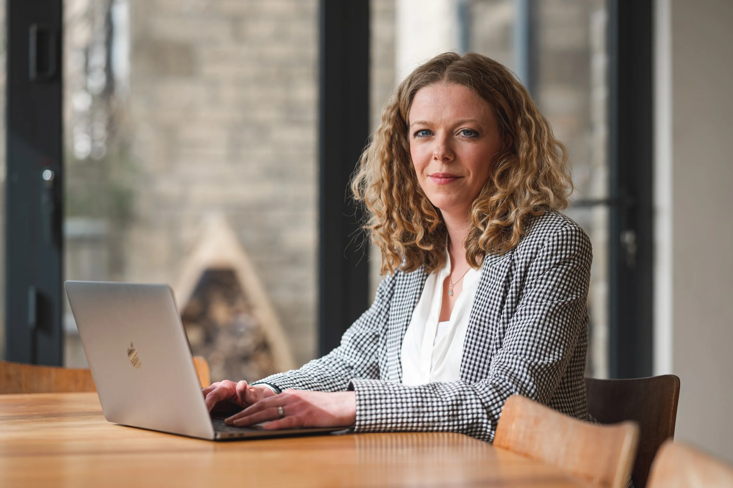 A woman with curly blonde hair wearing a checkered blazer and white blouse, sitting at a wooden table with a laptop in front of her, indoors with large windows in the background.
