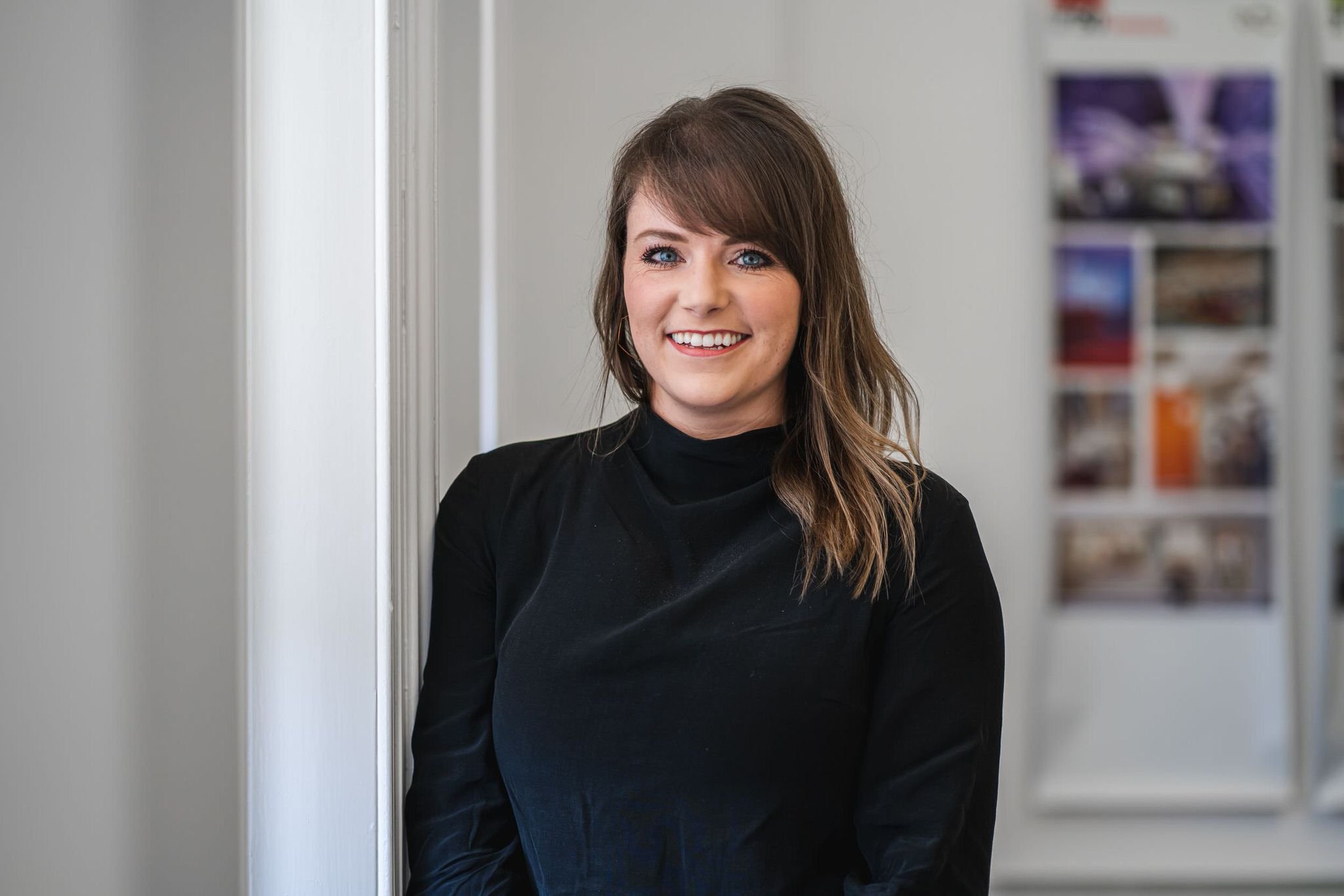 A young woman with shoulder-length brown hair and blue eyes, smiling and wearing a black top, standing indoors in front of a plain wall with some picture frames in the background.