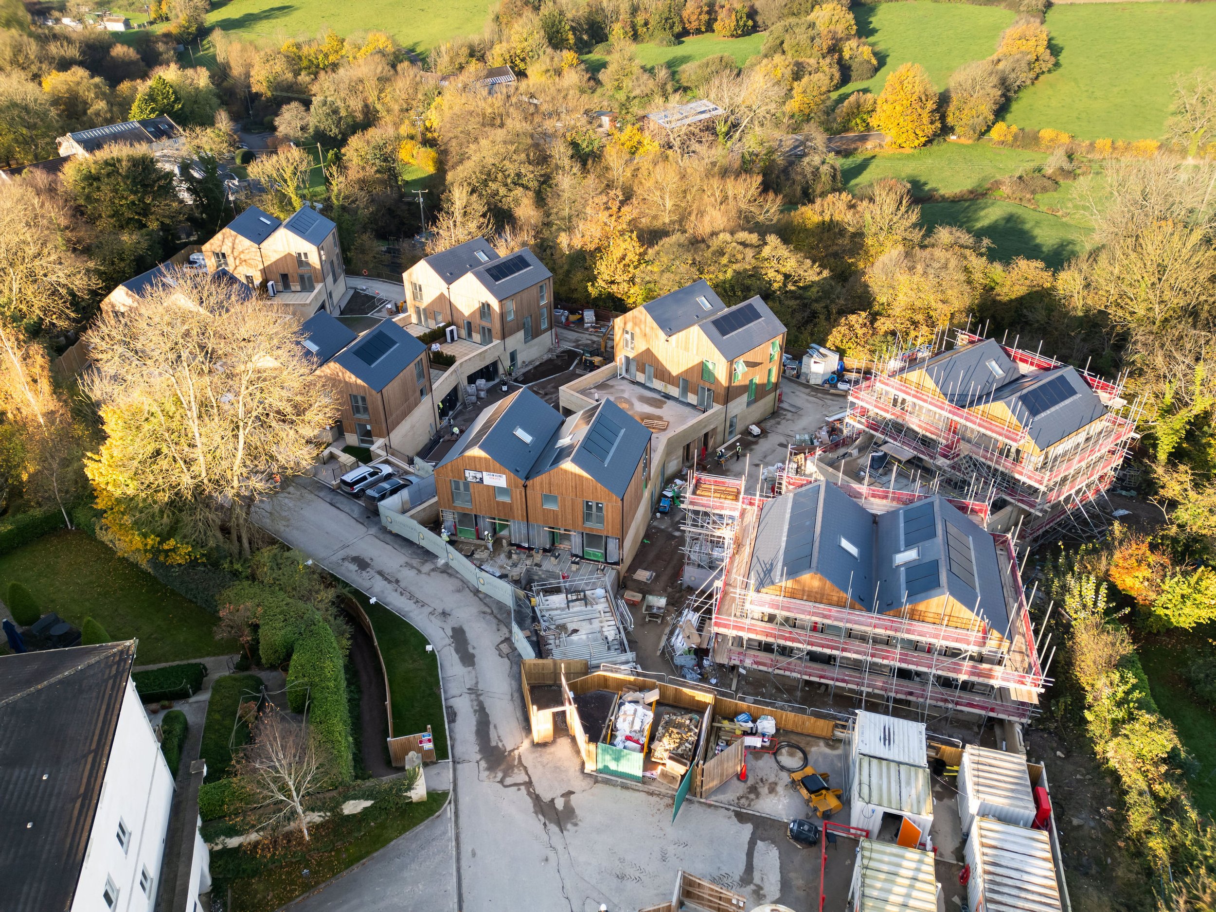 Aerial view of a housing development under construction with several completed houses and others being built with scaffolding, surrounded by trees and green fields.