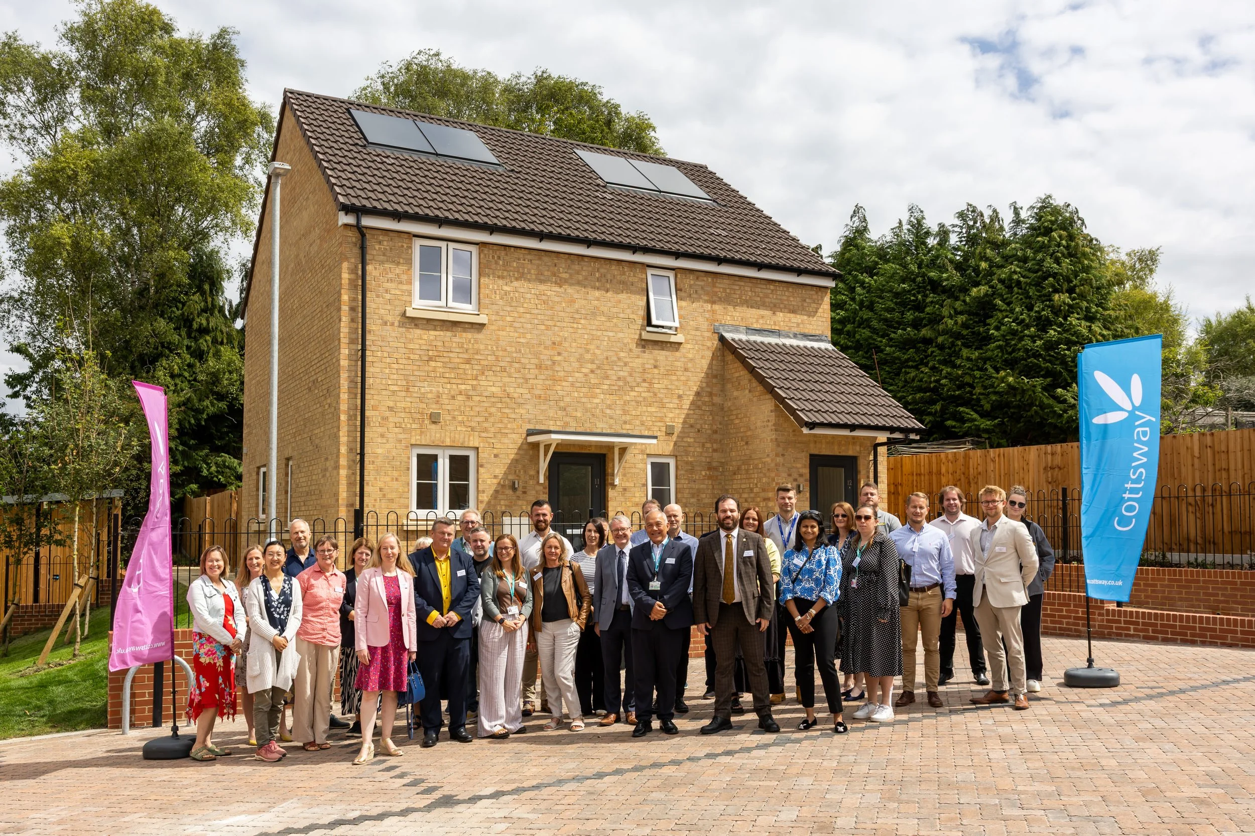 Group of people standing in front of a new brick residential building with solar panels on the roof, under a partly cloudy sky, during daytime. Two flags, one pink and one blue, with the blue flag displaying the word 'Cotswaby'.