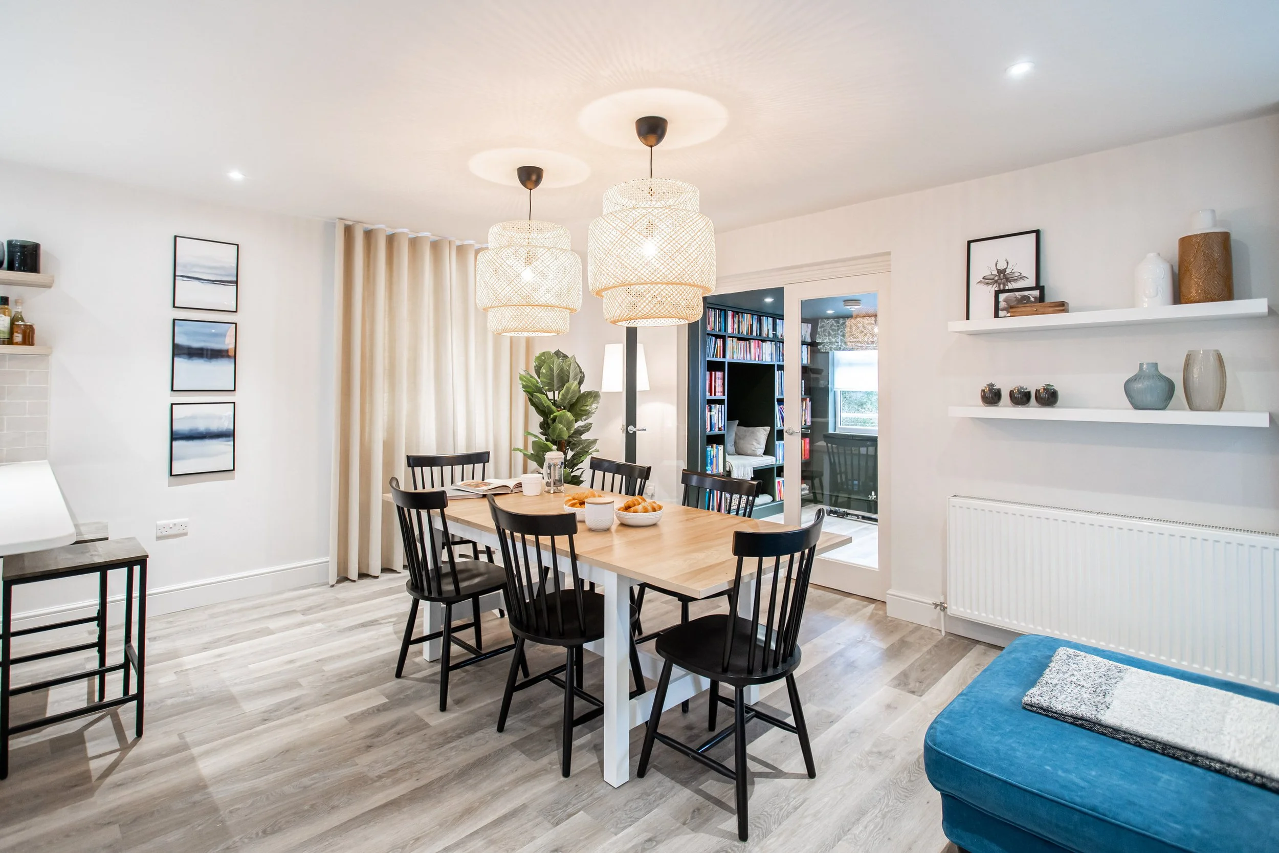 Bright dining room with a wooden table, six black chairs, three hanging woven pendant lights, a white radiator, built-in white shelves with decorative items, and a blue armchair. A doorway leads to a room with bookshelves.