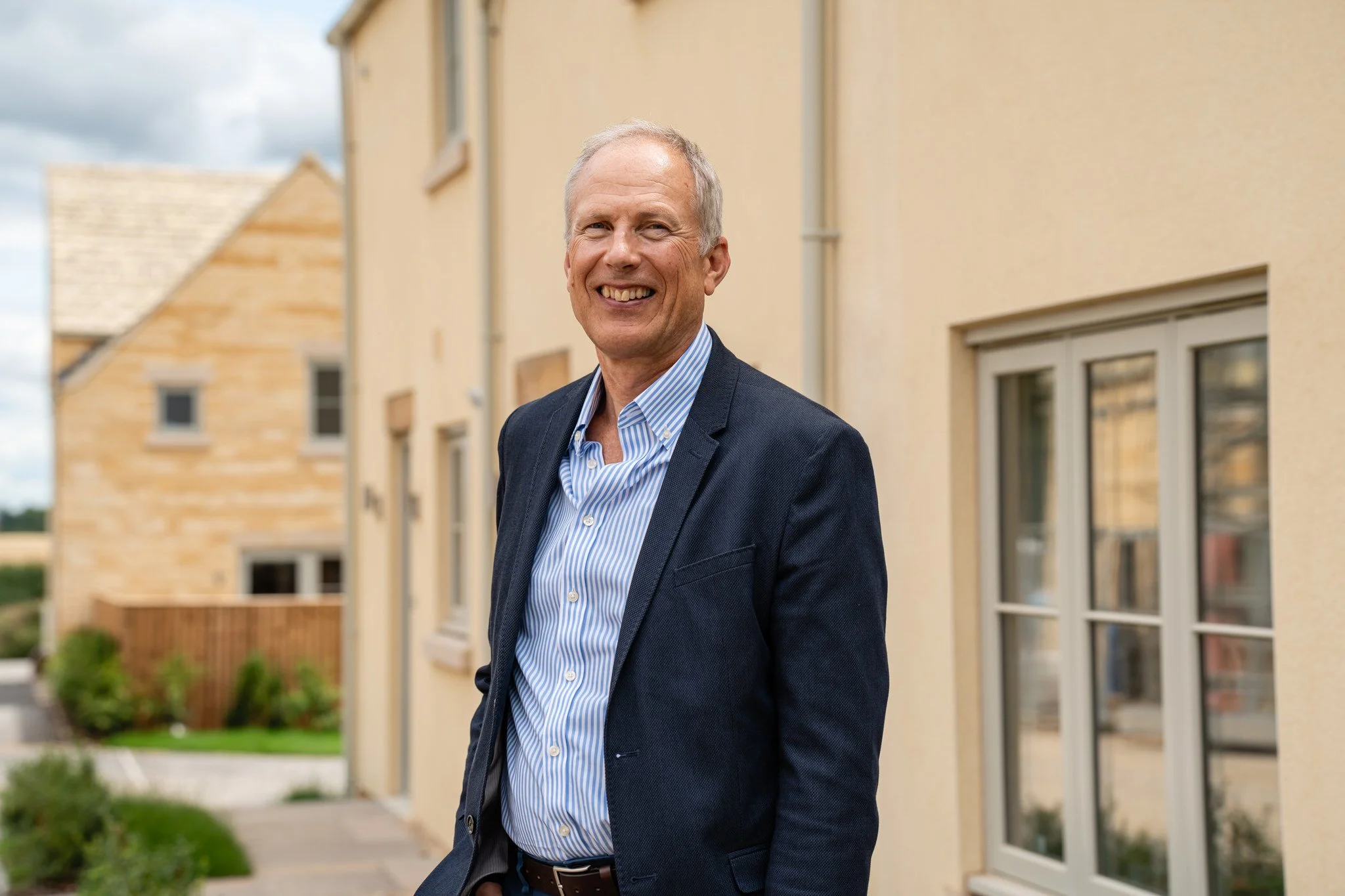 Smiling middle-aged man in a dark blazer and striped shirt standing outside near beige houses with windows and rooflines, with a garden in the background.