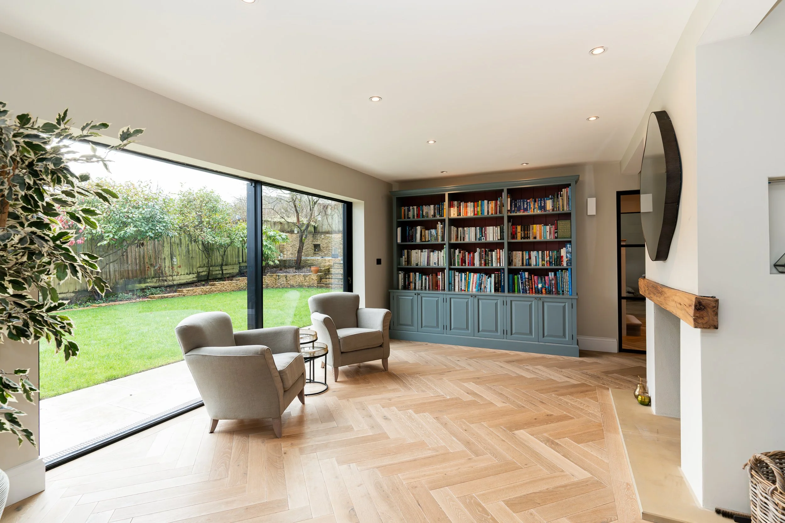 Living room with two gray armchairs and a glass side table near large sliding glass door leading to a green backyard, with a blue bookshelf filled with books against the wall, and a fireplace with a wooden mantel on the right.