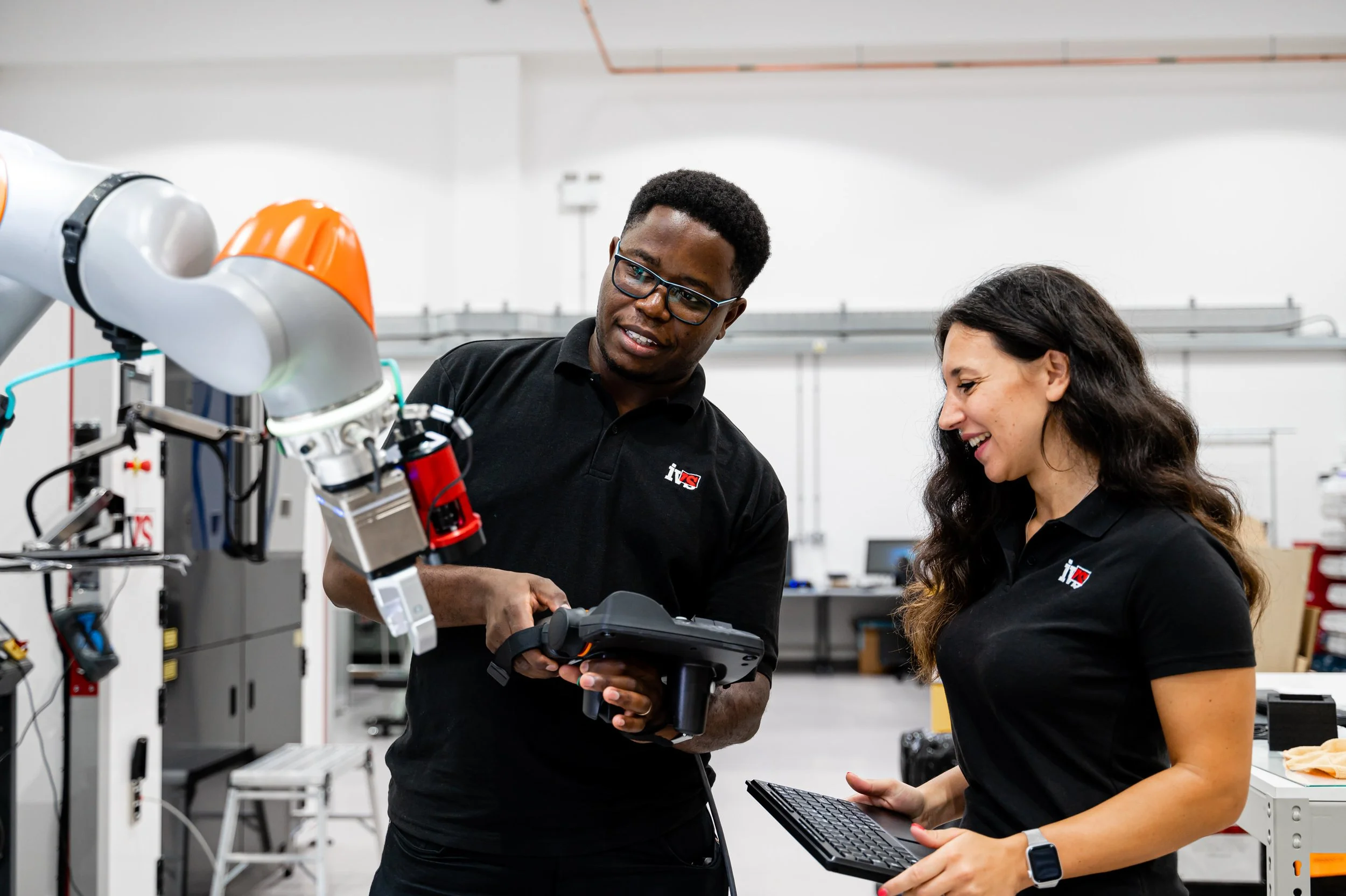 Two engineers in black shirts working with a robotic arm in a laboratory setting, discussing and operating a handheld device.