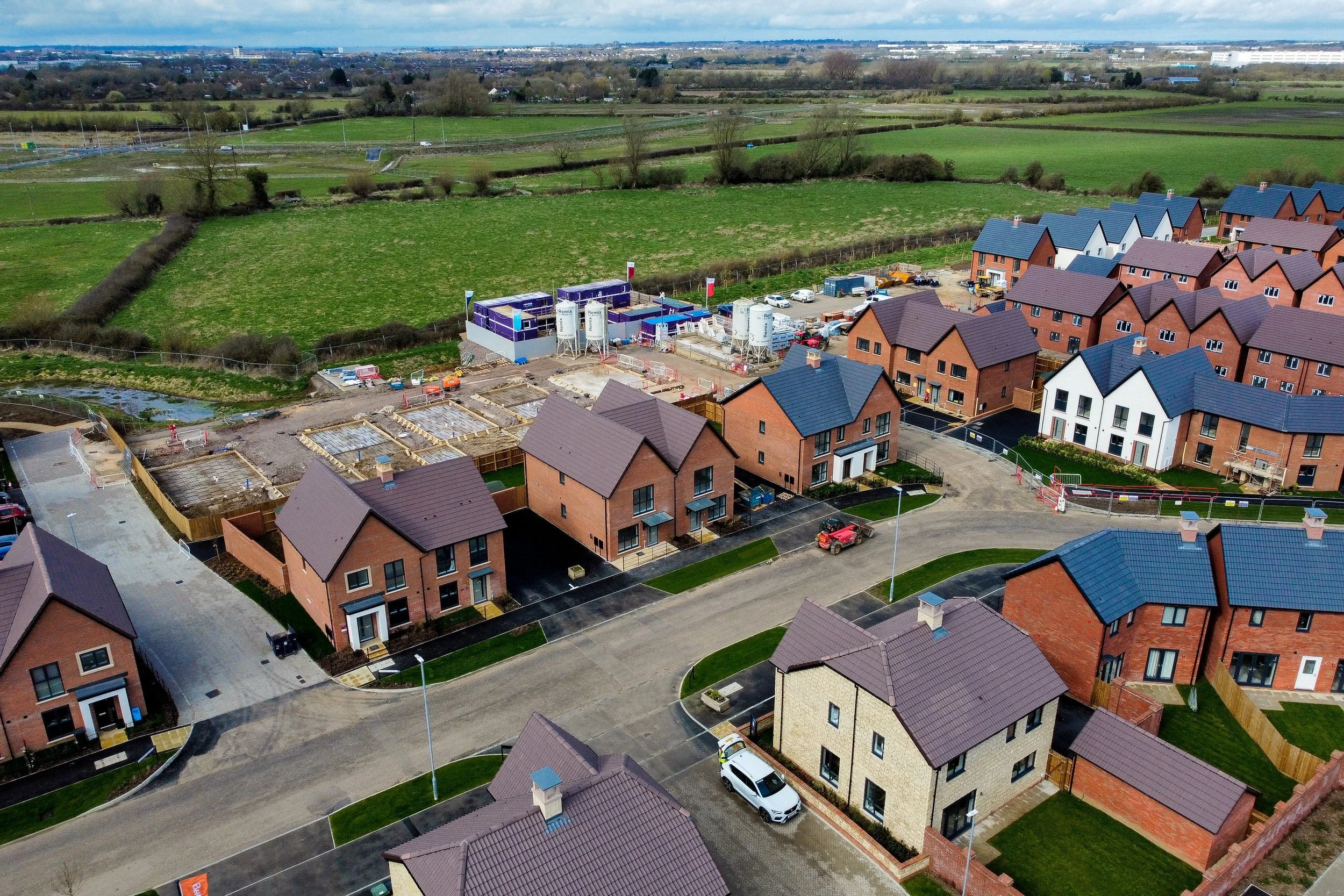 An aerial view of a new housing development with several houses, some under construction, and a construction site in a suburban area surrounded by green fields.