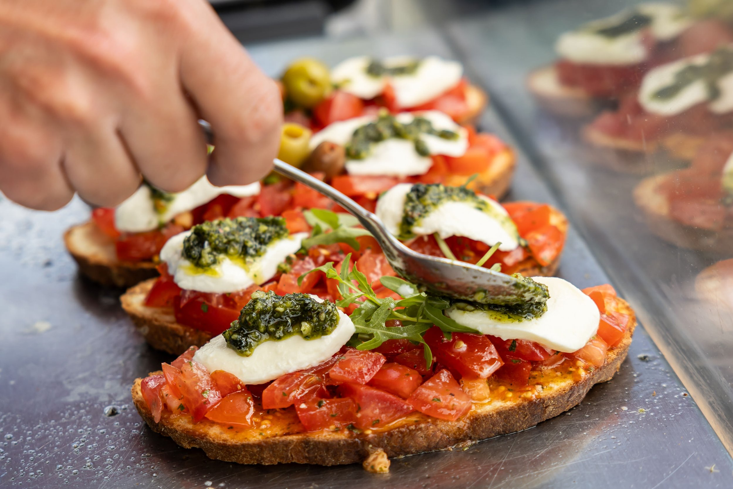 A person is using a spoon to add green sauce to small open-faced tomato and mozzarella sandwiches topped with green olives and fresh herbs on a metal tray.